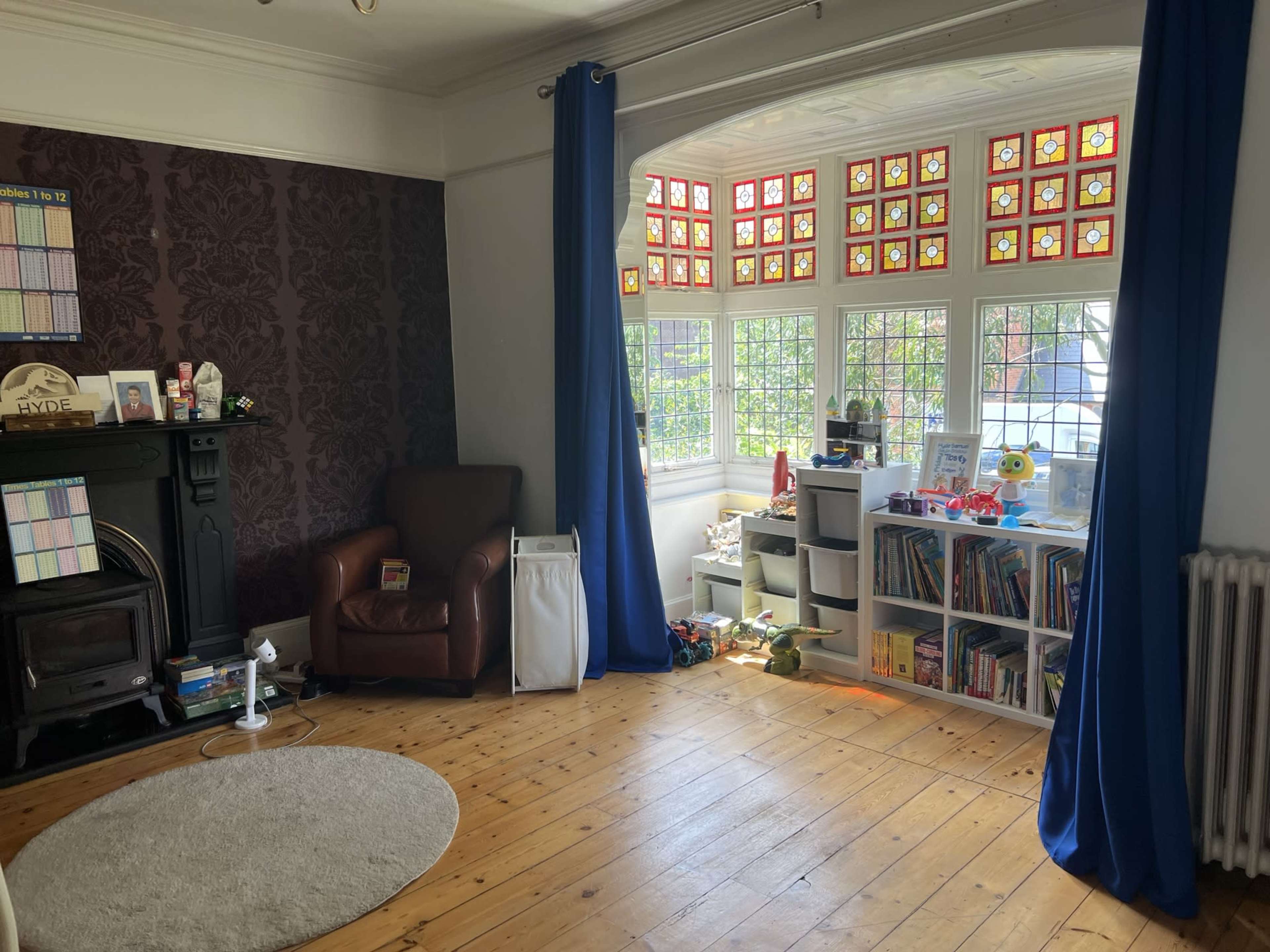 The room features a brown armchair, a fireplace, a light-colored rug, and a shelf filled with toys and books, under large windows adorned with colorful stained glass.