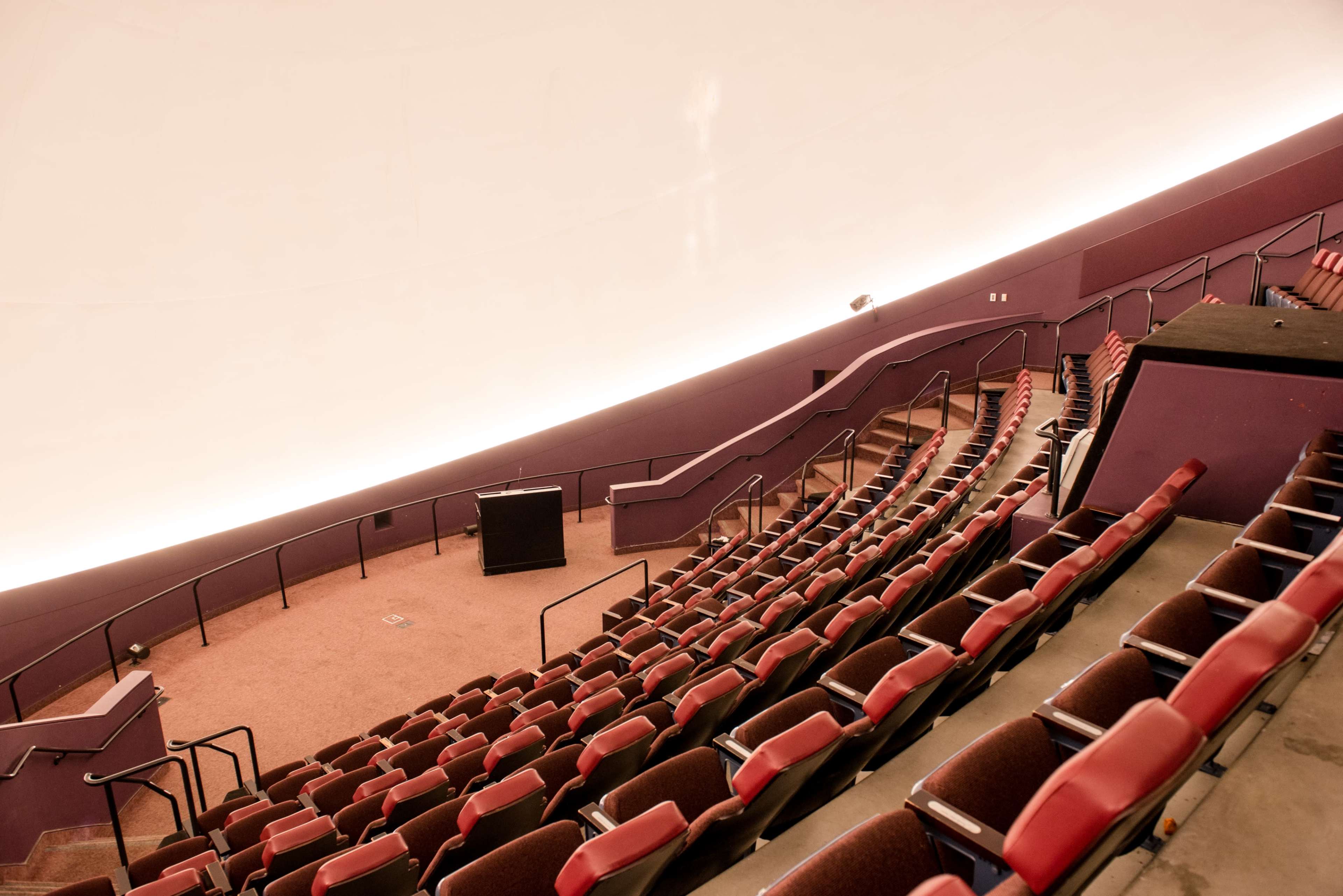 The image shows a partially empty auditorium with tiered seating and a large, curved ceiling.