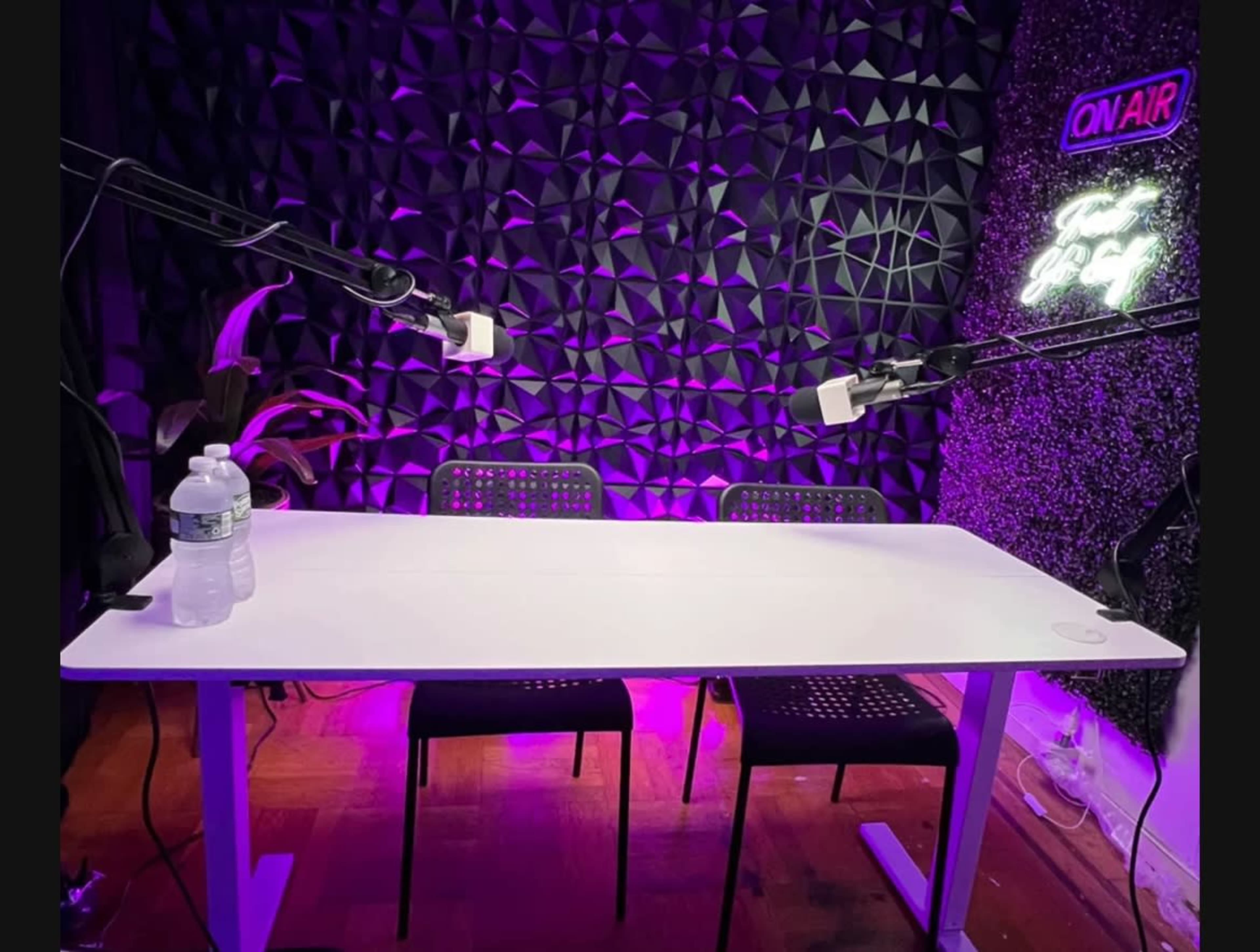 A white table with three black chairs and microphones is set up against a textured, illuminated wall featuring a neon "ON AIR" sign.