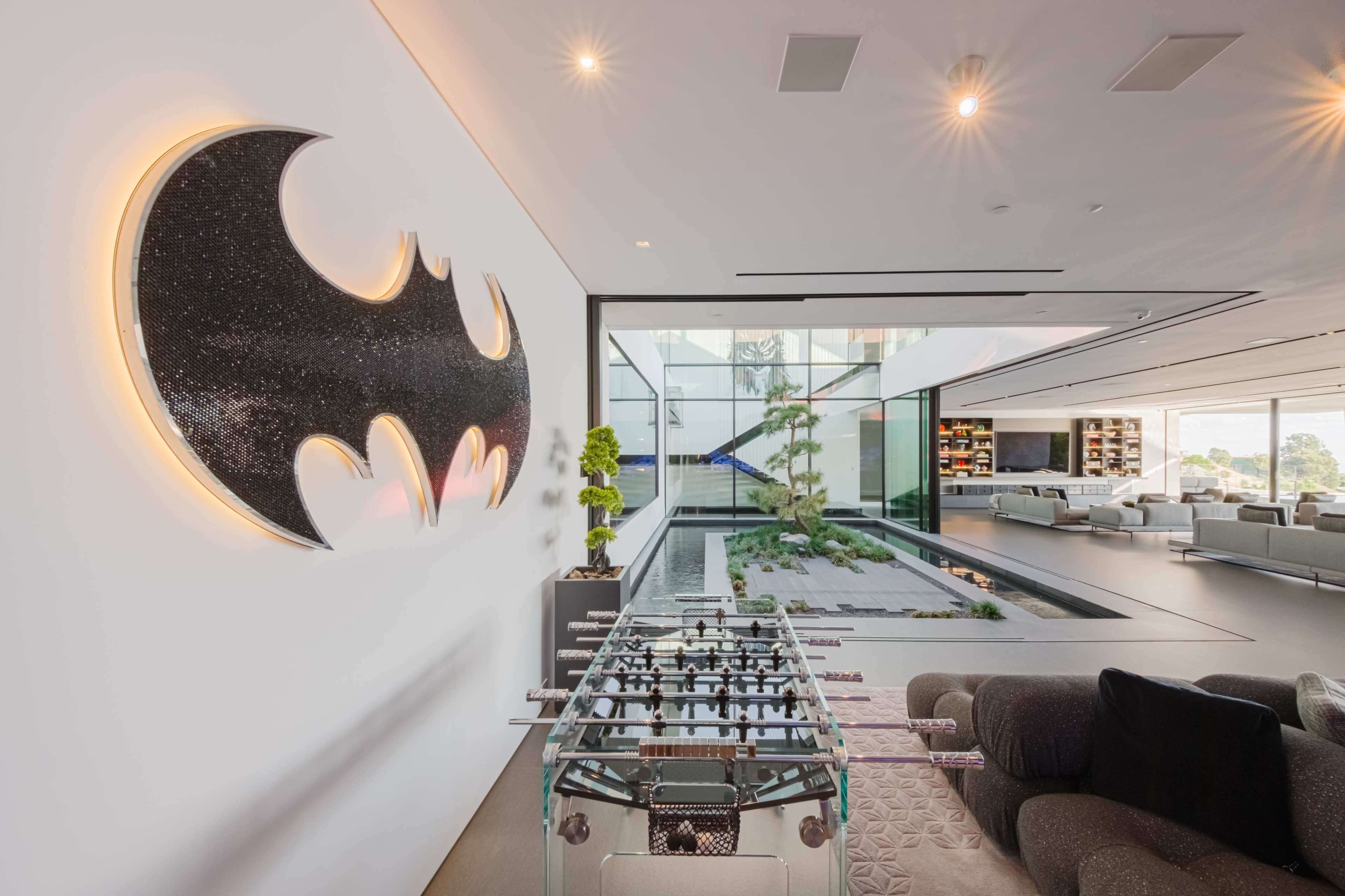 A modern living space features a black and gold Batman logo on the wall, with a glass wall revealing a serene indoor garden and a foosball table in the foreground.
