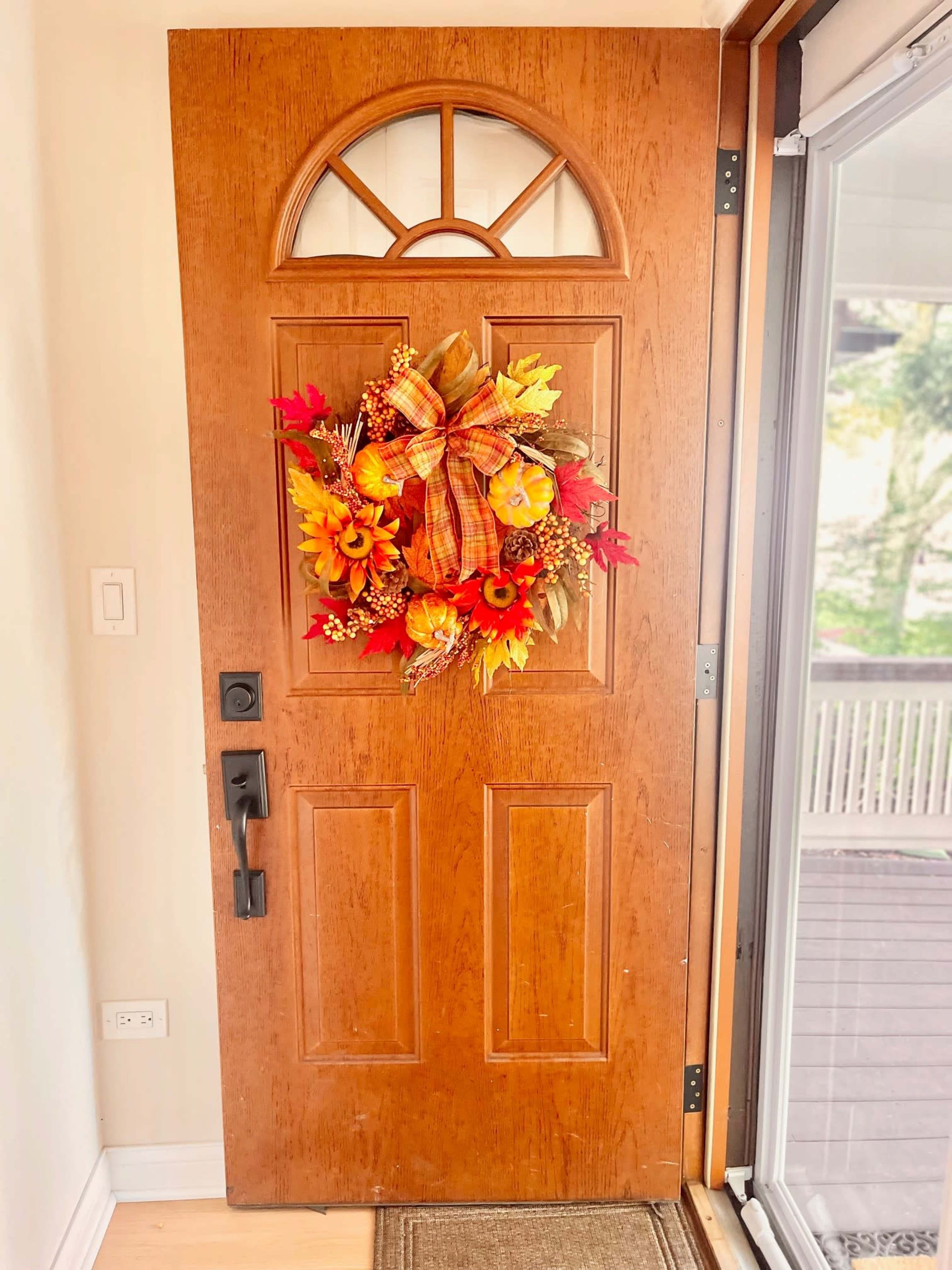 A wooden front door adorned with a colorful autumn wreath featuring orange and yellow flowers, along with decorative leaves and a bow.