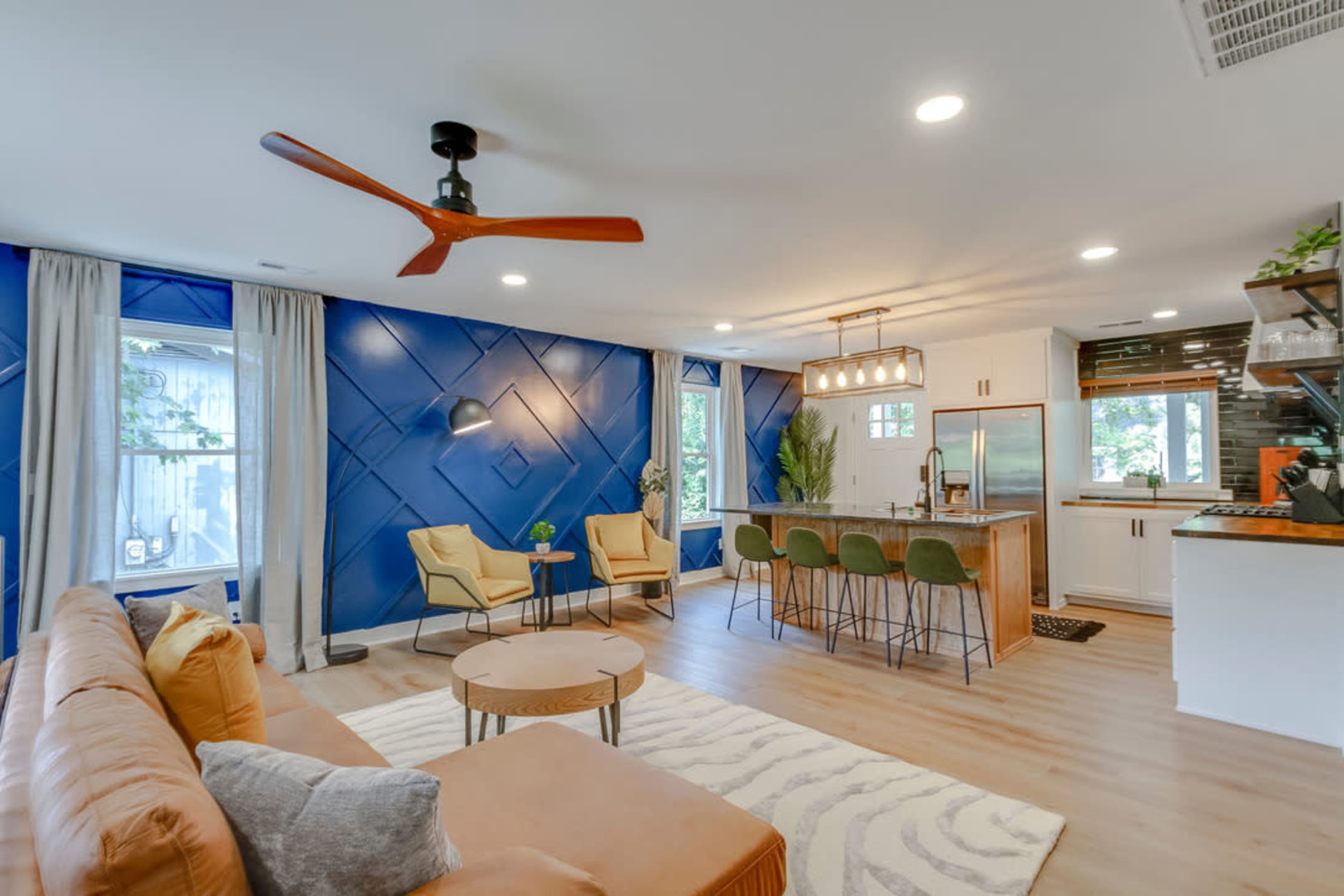The image shows a modern living room with a blue accent wall, a brown sofa, two yellow chairs, and a kitchen area with bar stools.