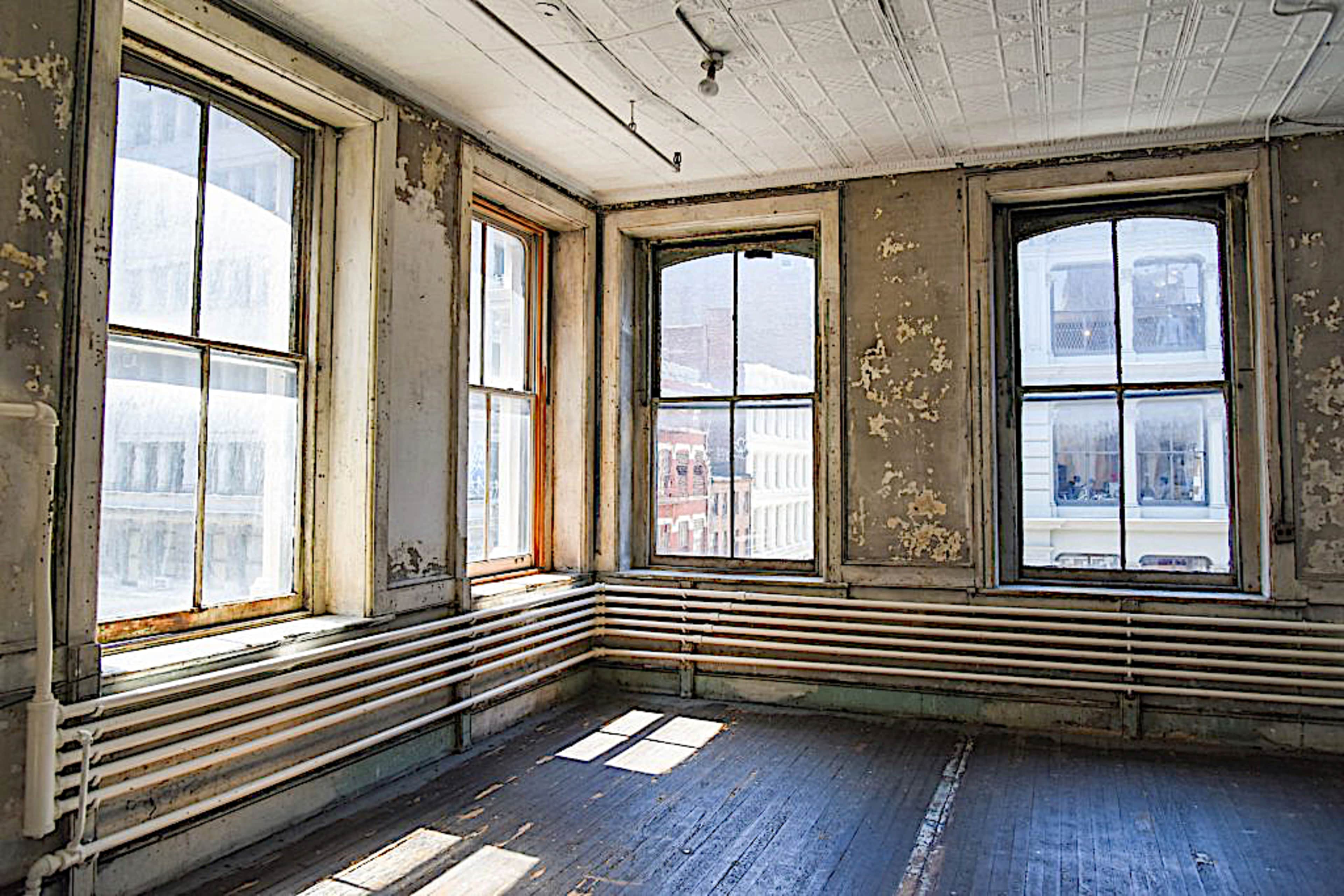 An empty room with large, peeling windows and wooden floors, illuminated by sunlight streaming through the glass.