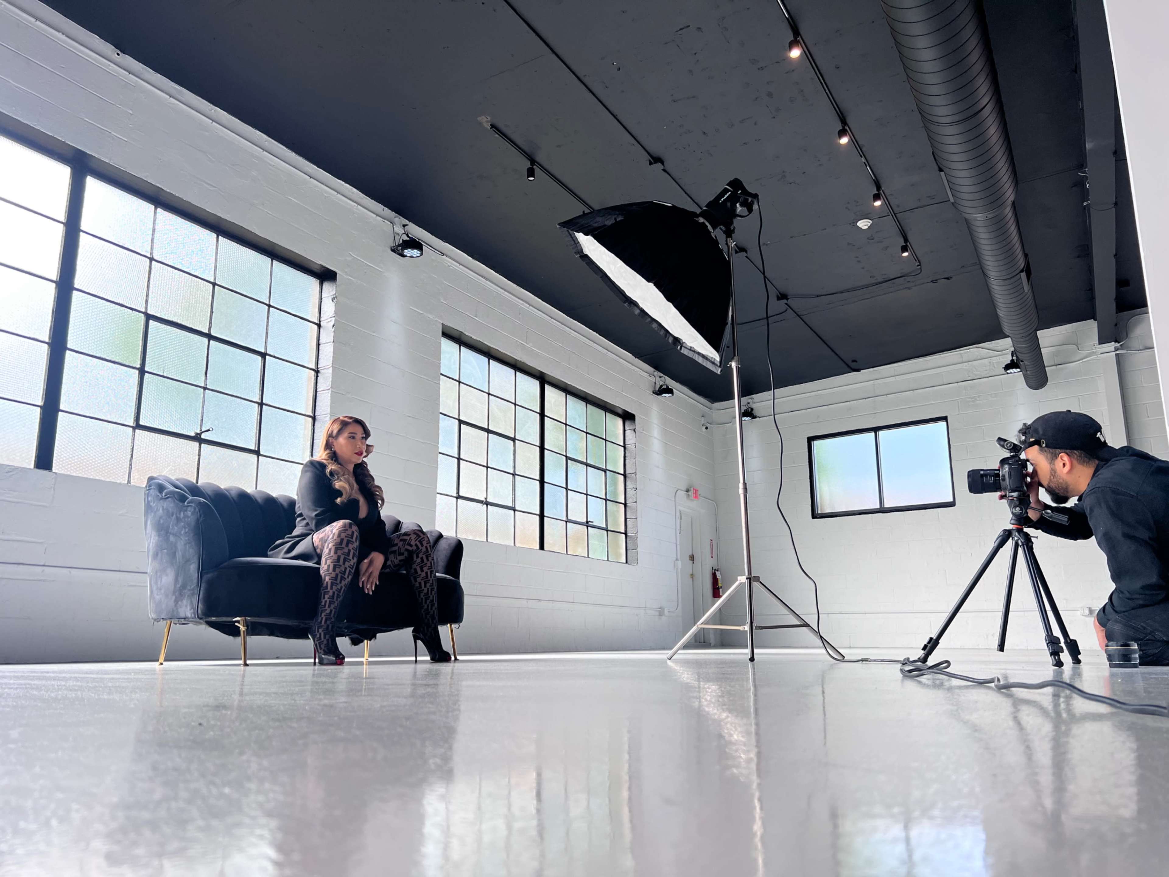 A photographer captures a model sitting on a black couch in a large, well-lit studio space.