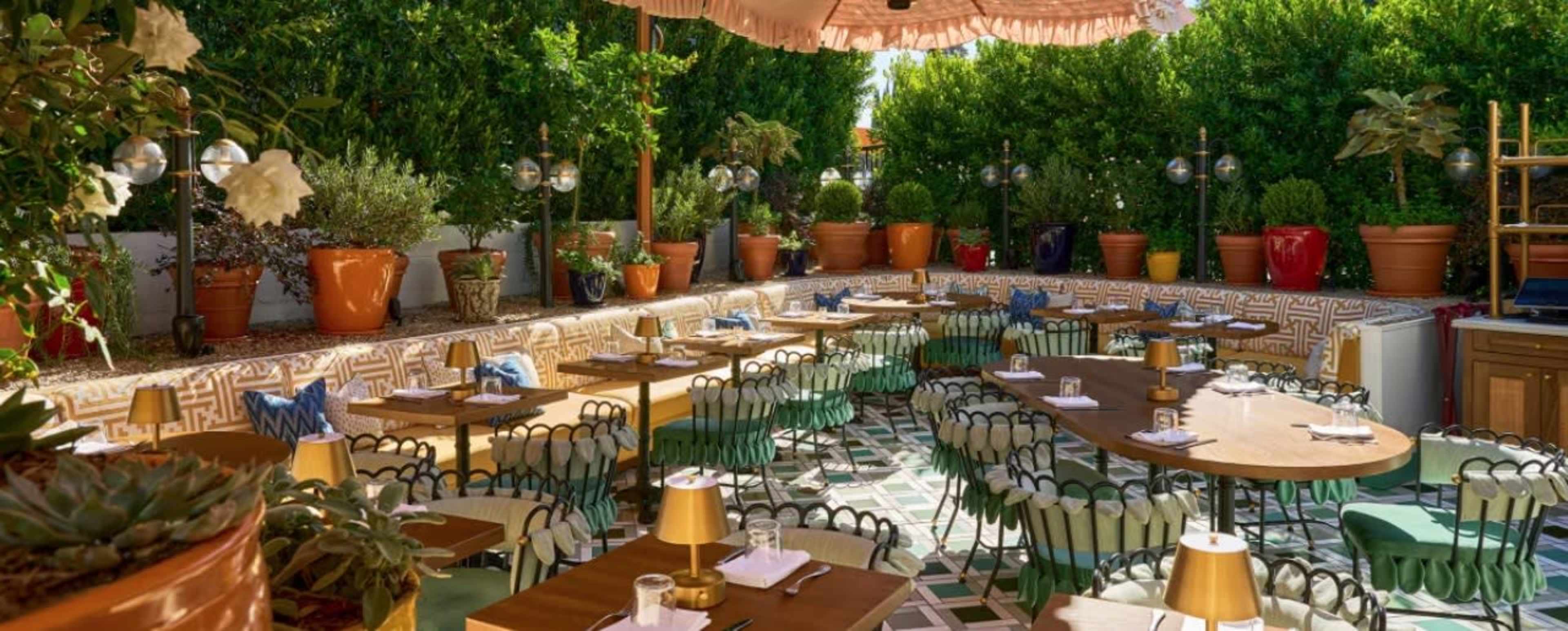 The image shows an outdoor restaurant setting with tables and chairs arranged under a large umbrella, surrounded by potted plants and greenery.