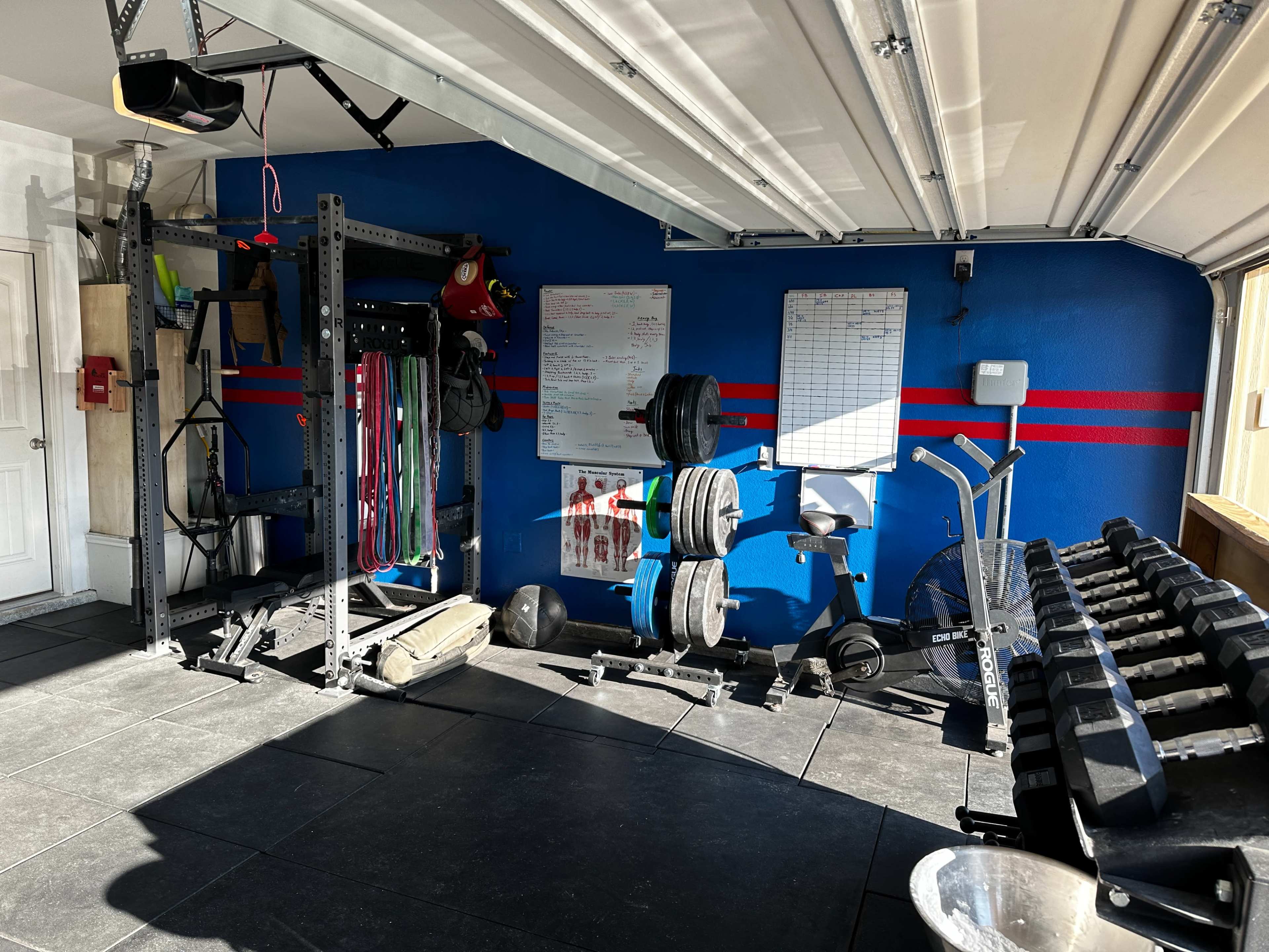 A home gym setup in a garage, featuring weightlifting equipment, exercise bands, and fitness charts on the walls.
