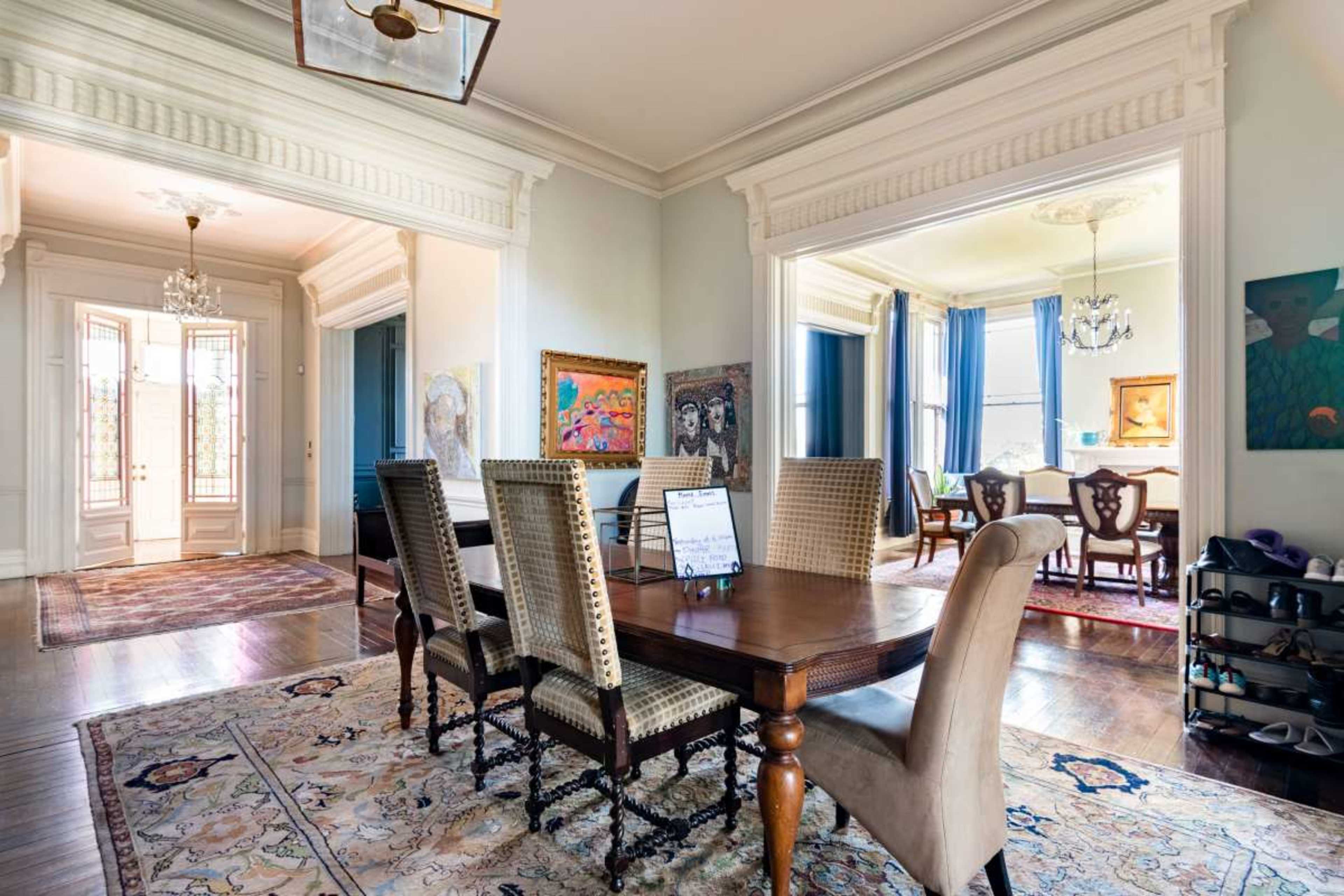 The image shows a spacious dining area featuring a wooden table surrounded by upholstered chairs, with doorways leading to additional rooms adorned with artwork and large windows.