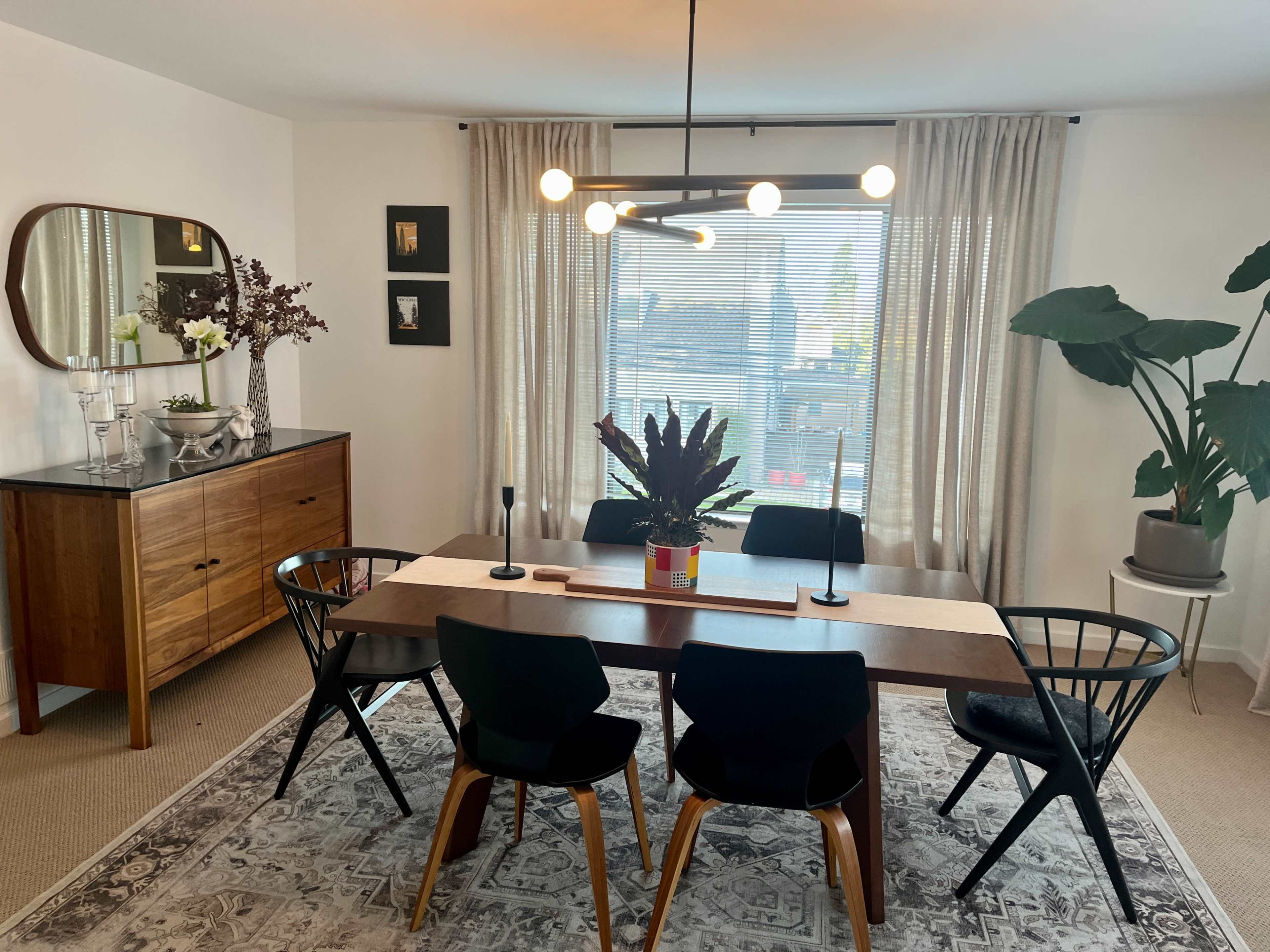 A dining area with a wooden table surrounded by black chairs, a large window with sheer curtains, and a sideboard with decorative items.