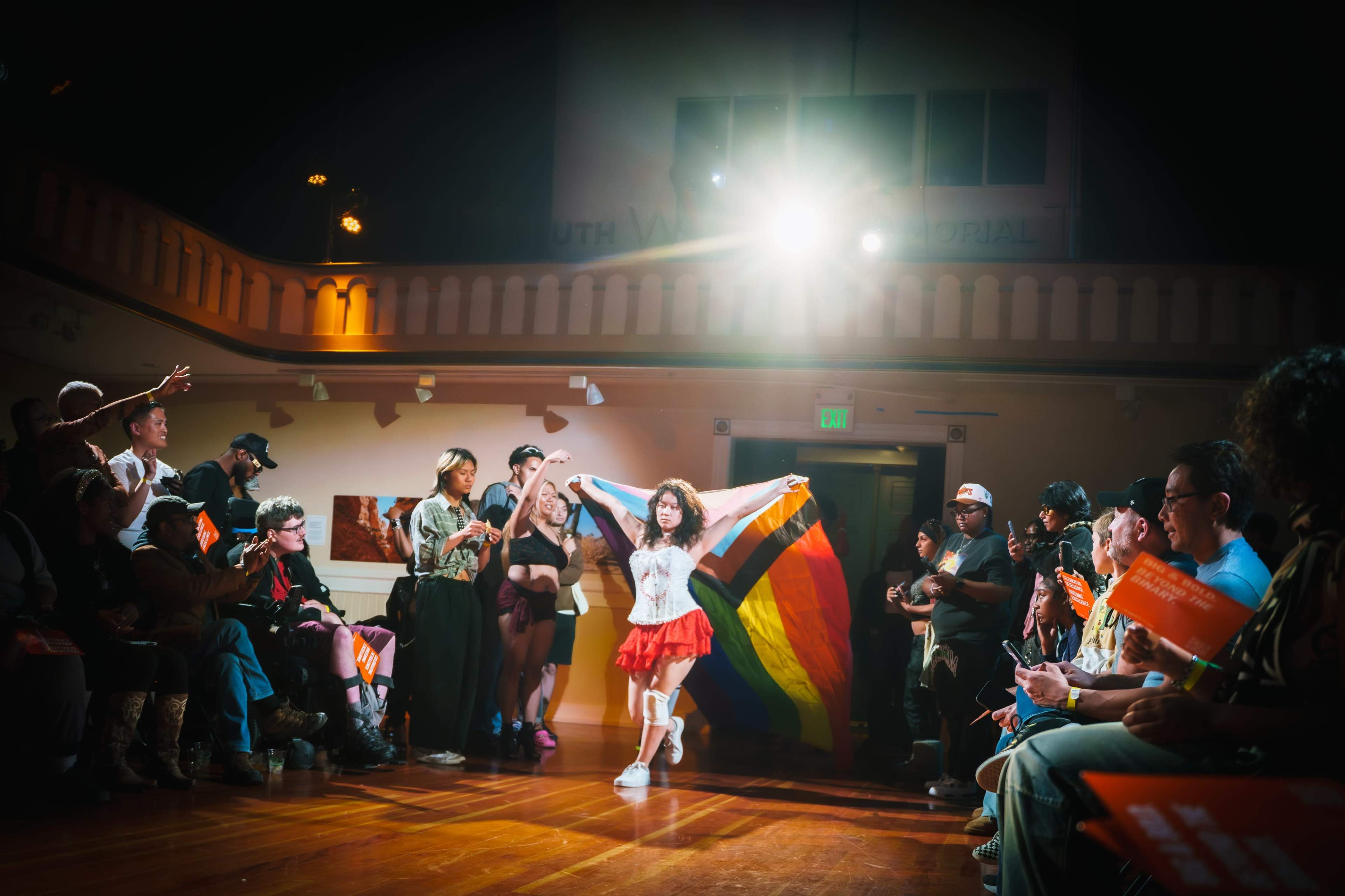 A performer holds a large rainbow flag while dancing on a runway in front of an audience.