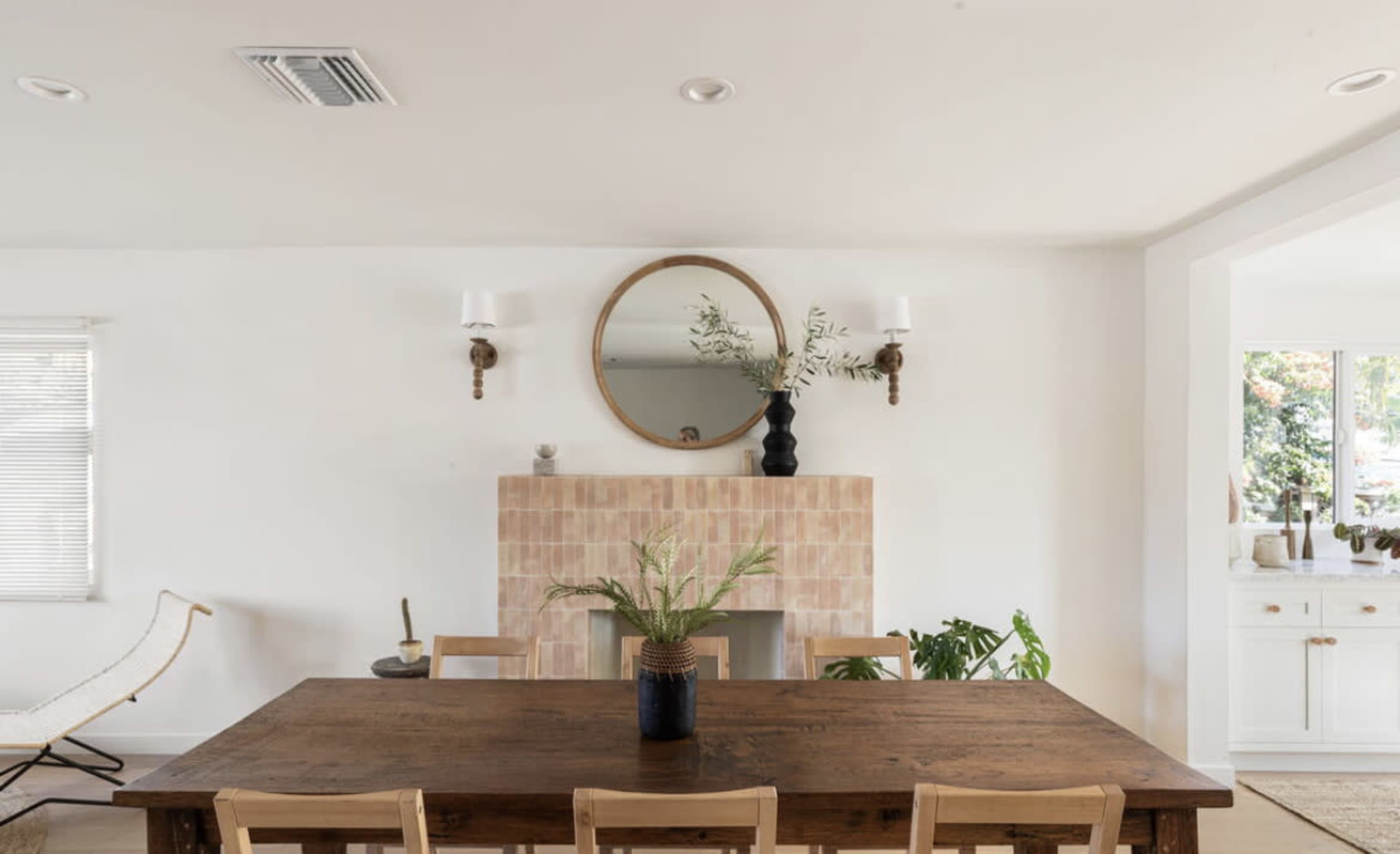 A modern dining area features a wooden table with light-colored chairs, a simple vase with plants, a round mirror above a tiled fireplace, and wall sconces.