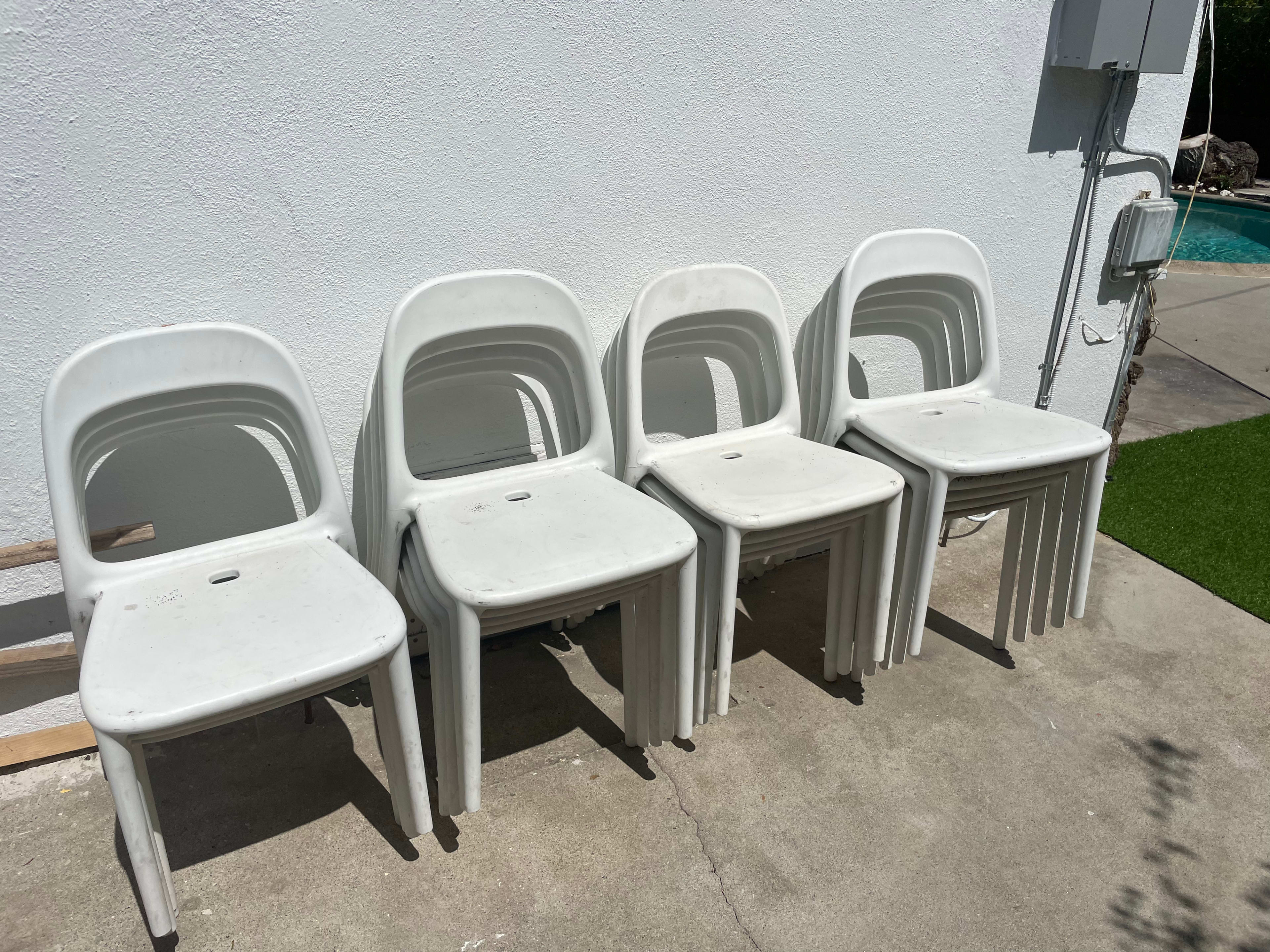 Four white plastic chairs are stacked against a white wall on a concrete surface.
