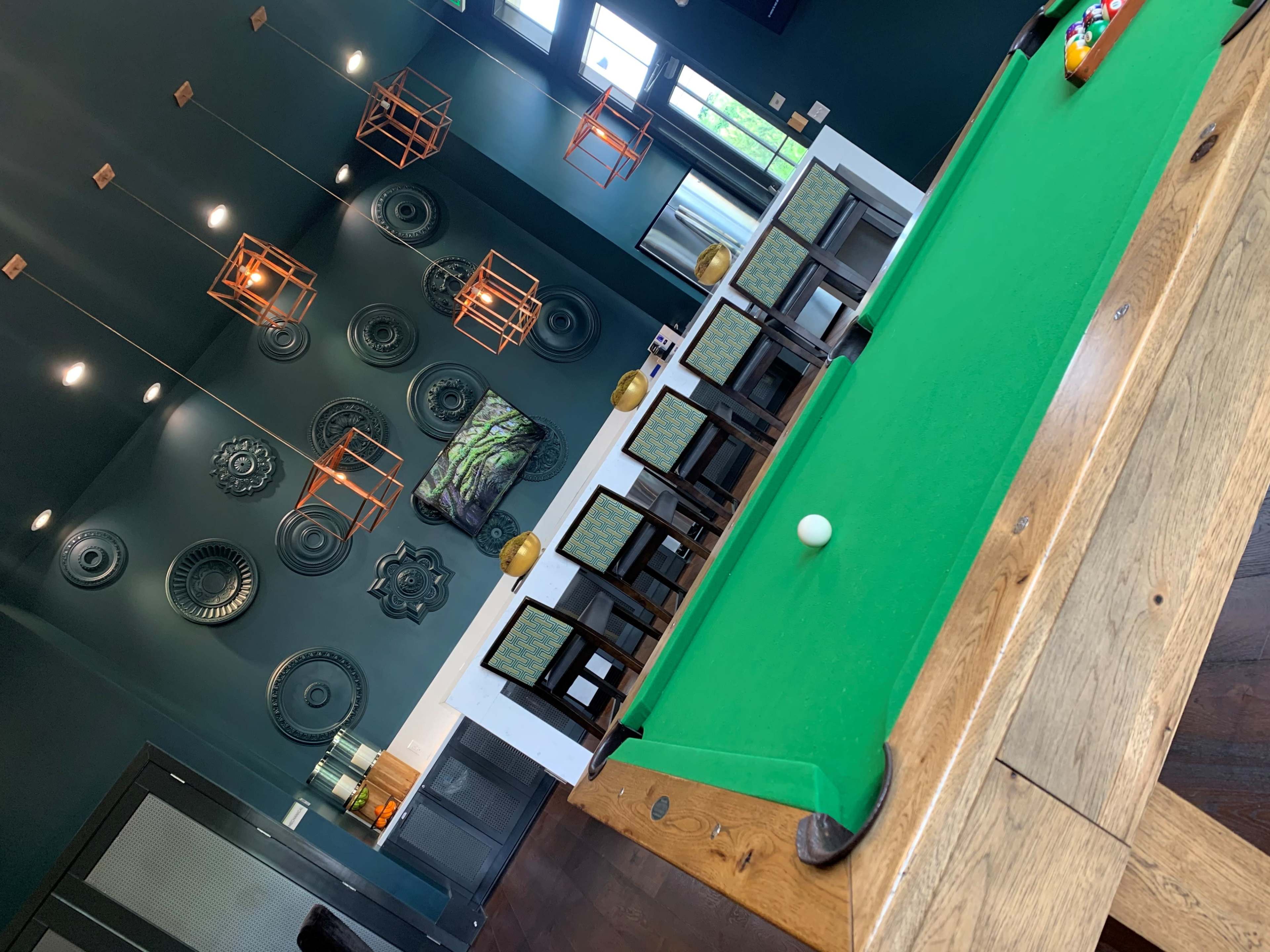 A billiards table with a white cue ball sits in a room featuring patterned wall art and hanging light fixtures.