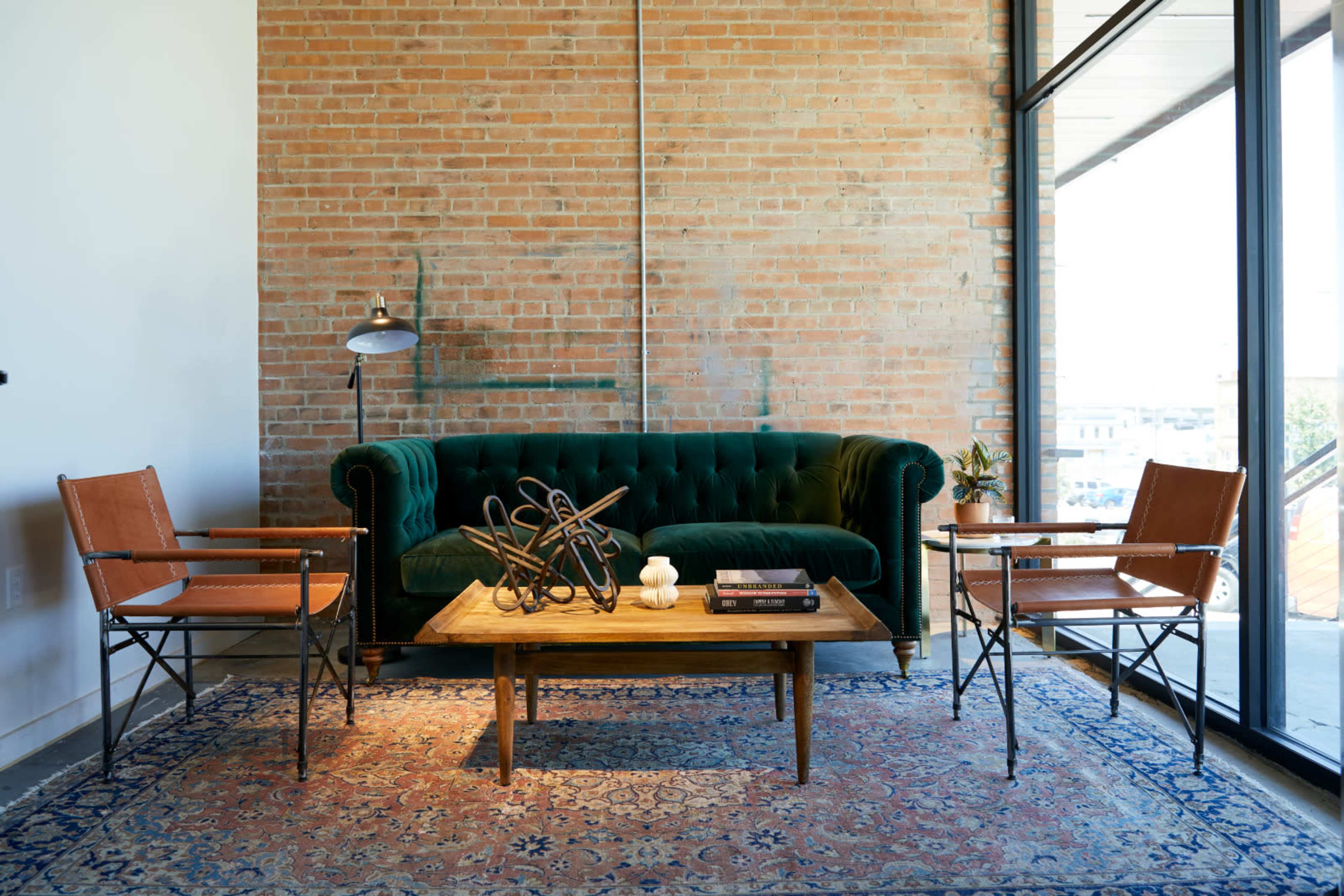 A green velvet couch is placed against a brick wall, accompanied by two leather chairs and a wooden coffee table on a patterned rug.