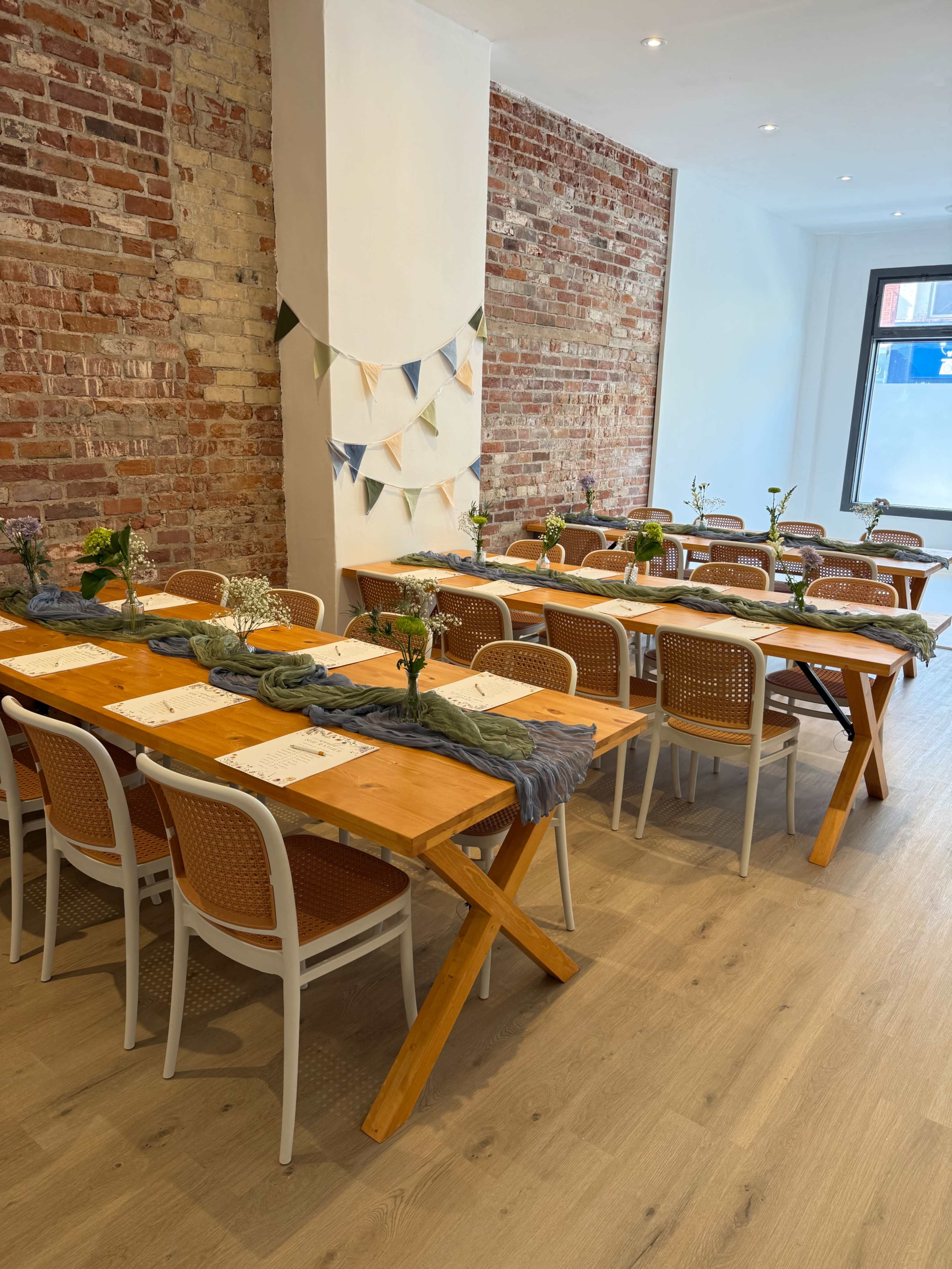 The image shows a dining area with wooden tables arranged neatly, decorated with floral centerpieces and bunting along the brick wall.