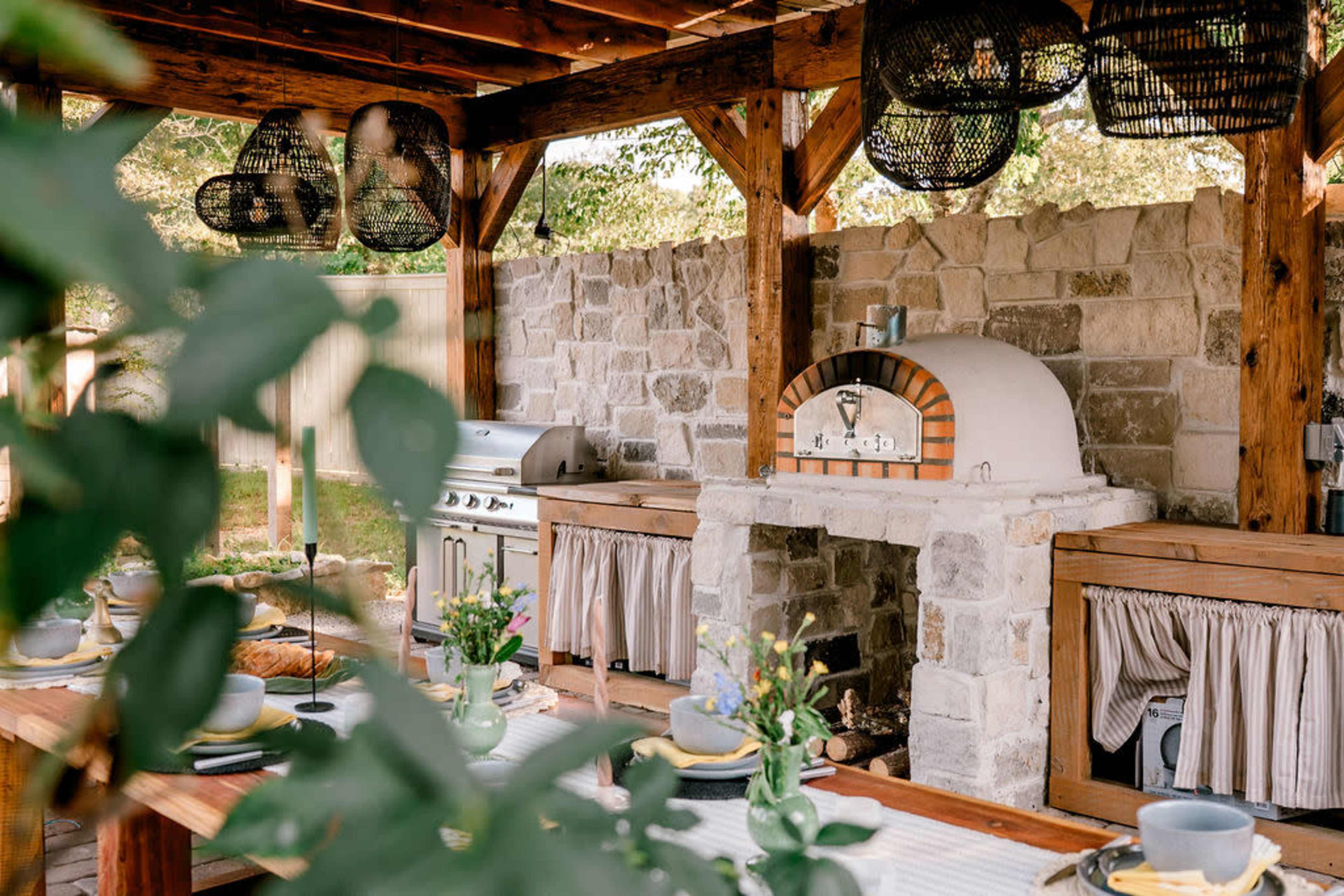 The image shows an outdoor kitchen area featuring a stone pizza oven, a grill, and a dining table set with dishes and floral arrangements under a wooden pergola.
