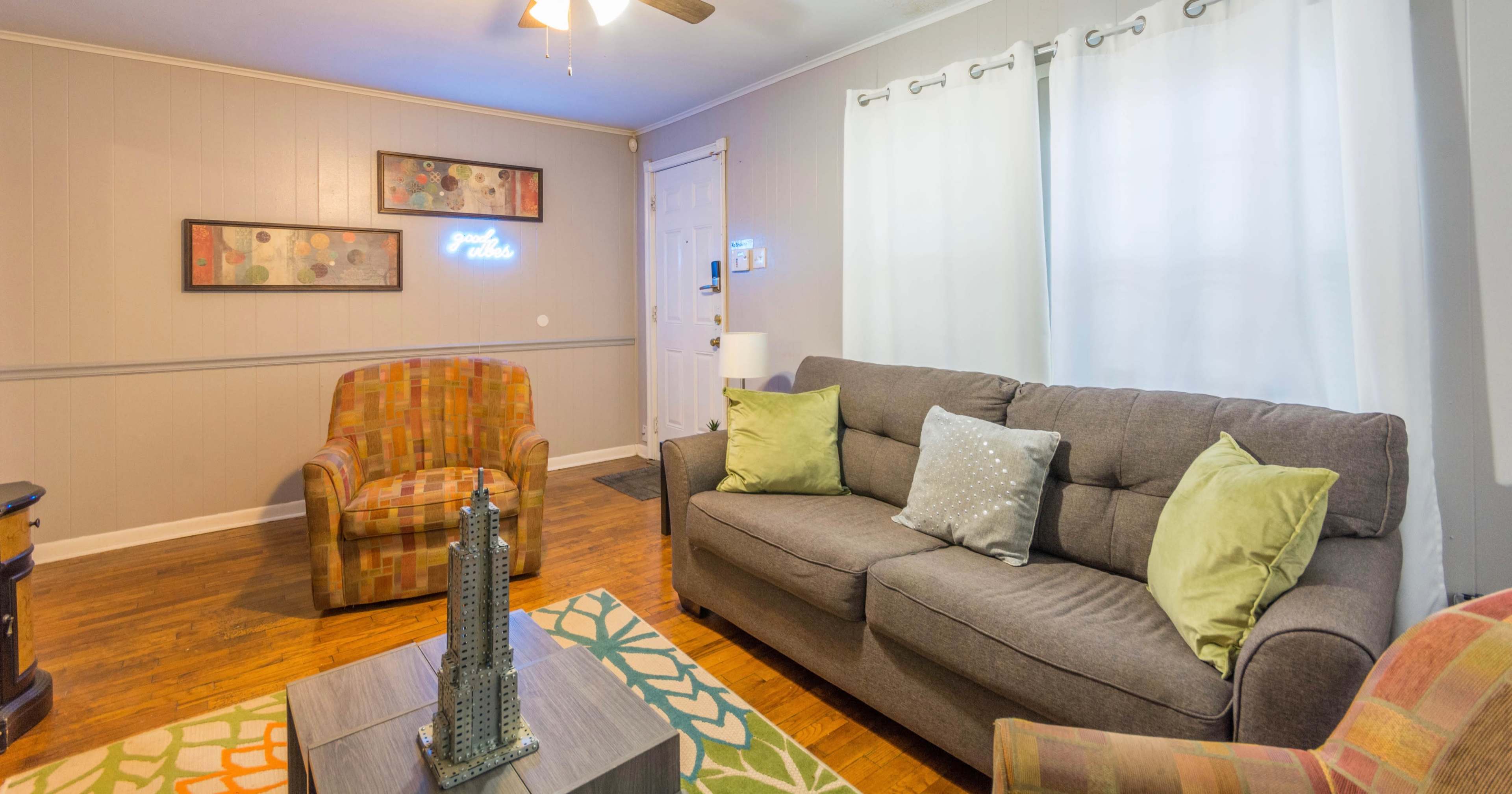 The living room features a gray sofa with decorative pillows, a patterned area rug, and two armchairs, along with a coffee table and wall art.