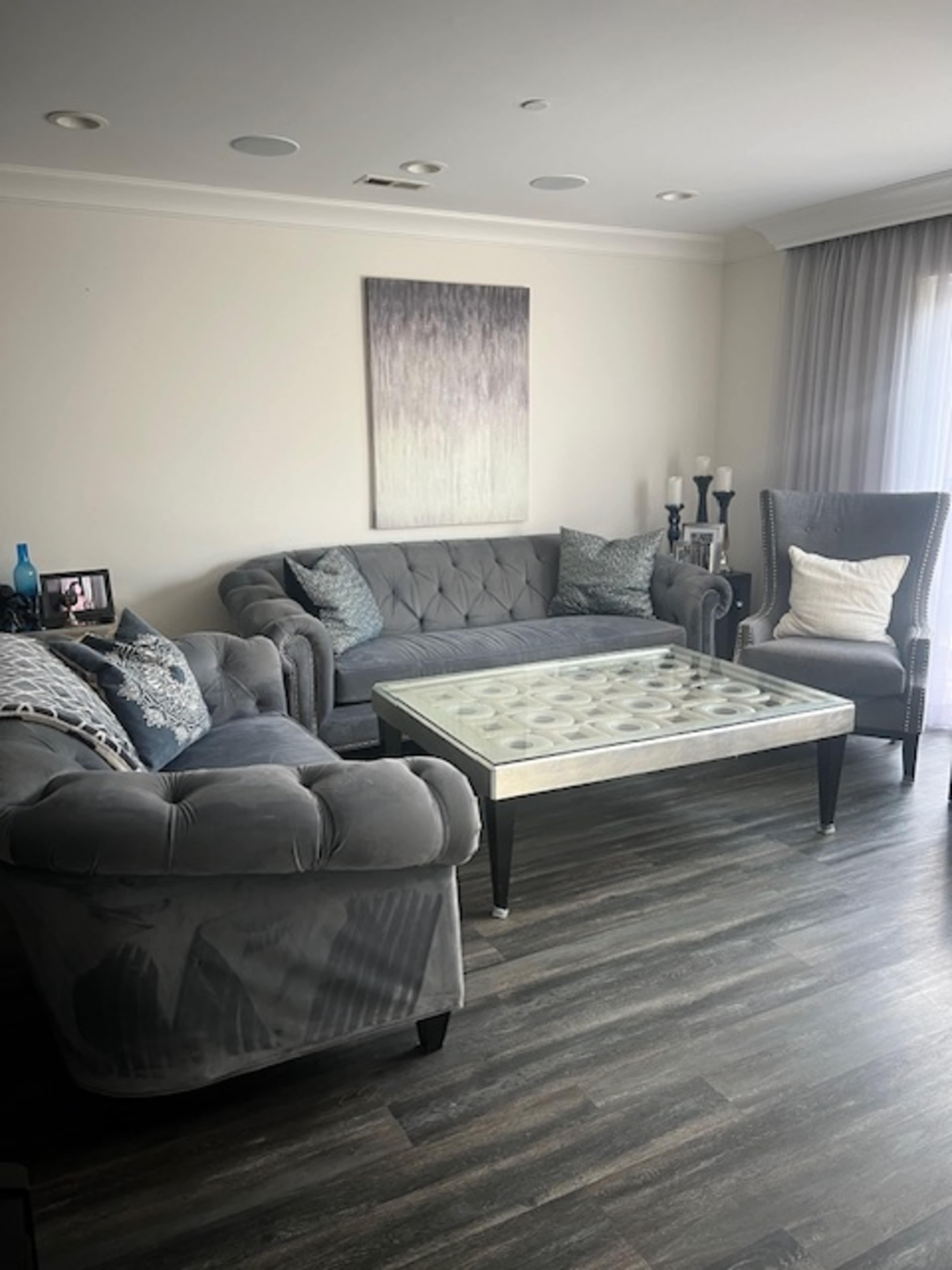 The image shows a modern living room with a gray tufted sofa, armchair, and a glass-top coffee table centered on a wooden floor.
