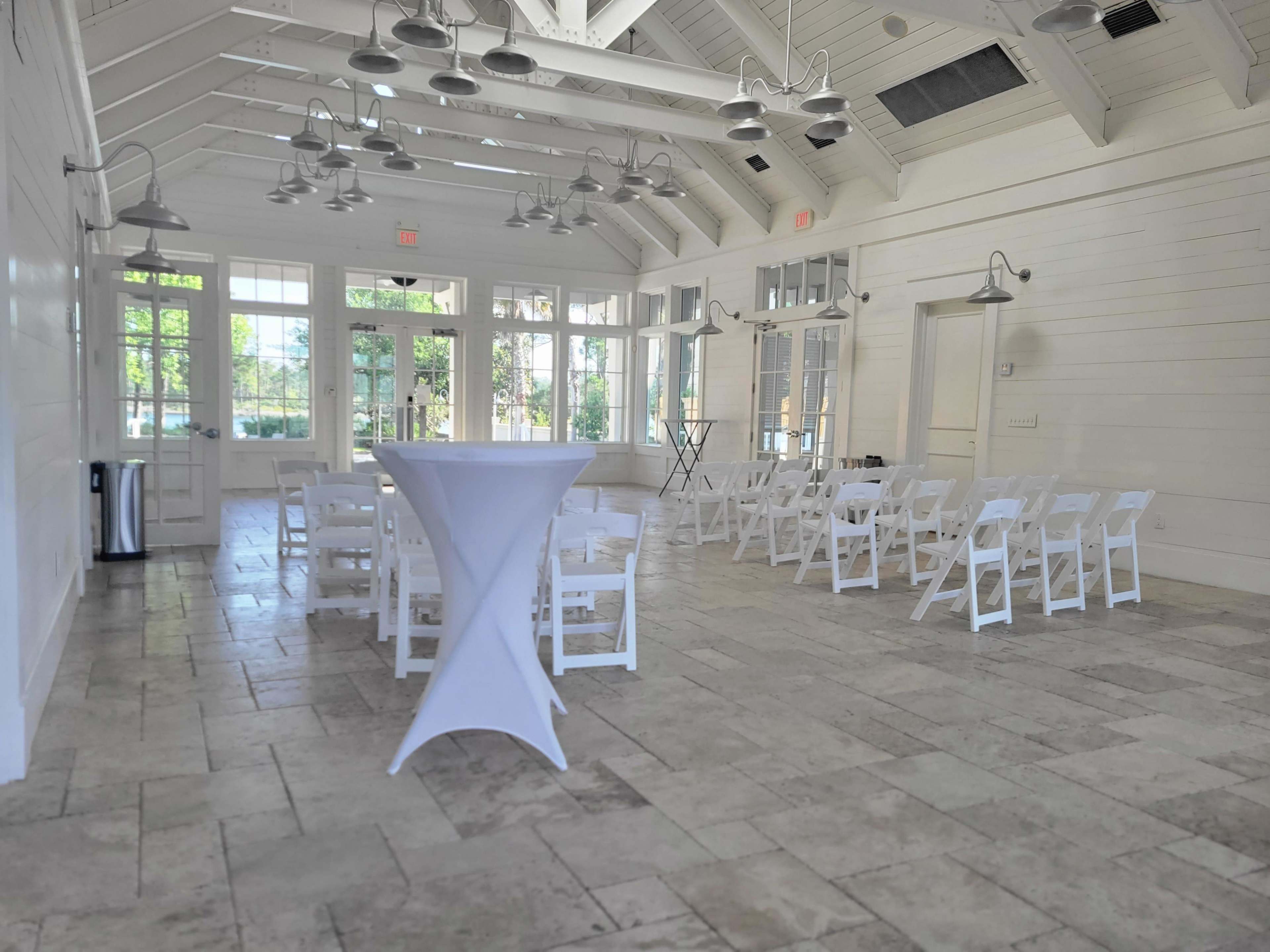 A bright, spacious event room with white walls, a tiled floor, and rows of white chairs arranged in front of a tall table.