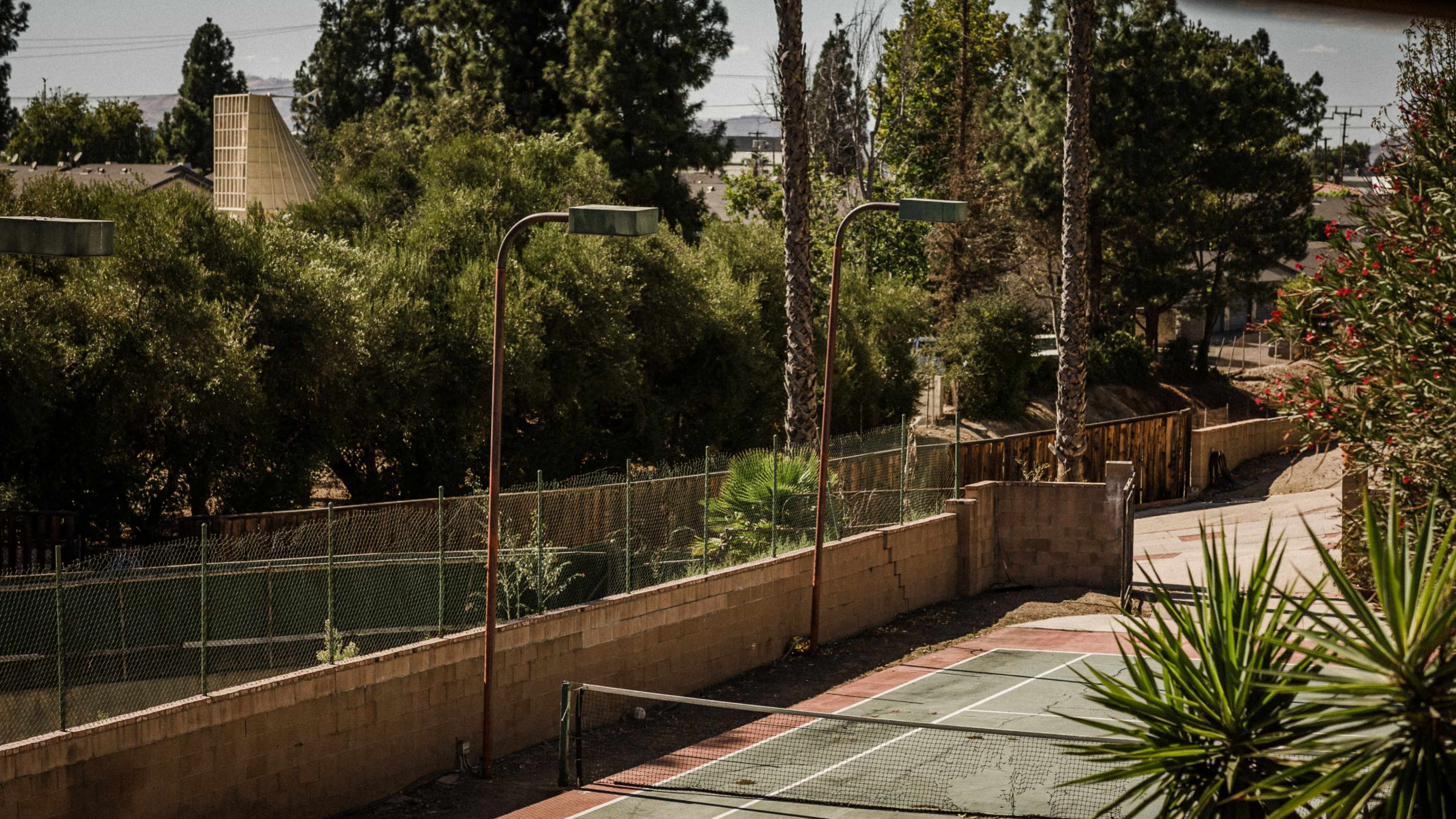 A tennis court bordered by trees and lighting fixtures, set against a backdrop of a residential area.