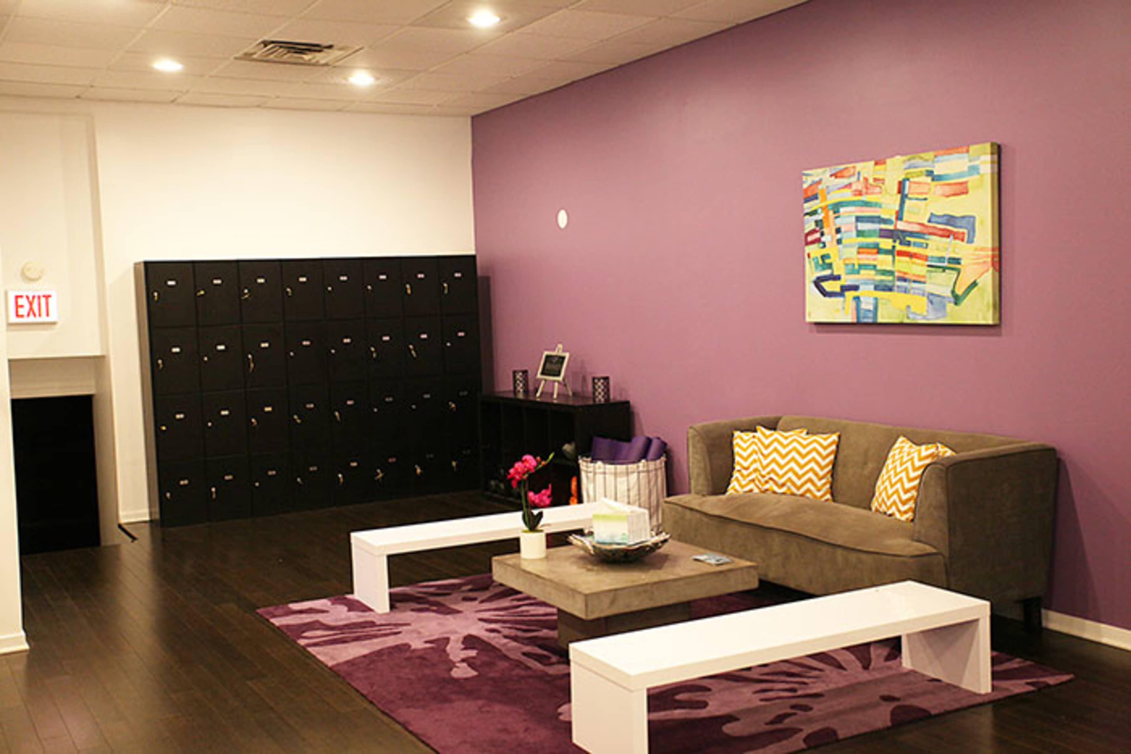 The image shows a modern office space featuring a purple accent wall, a gray sofa with patterned cushions, a coffee table, and a row of black lockers against the wall.
