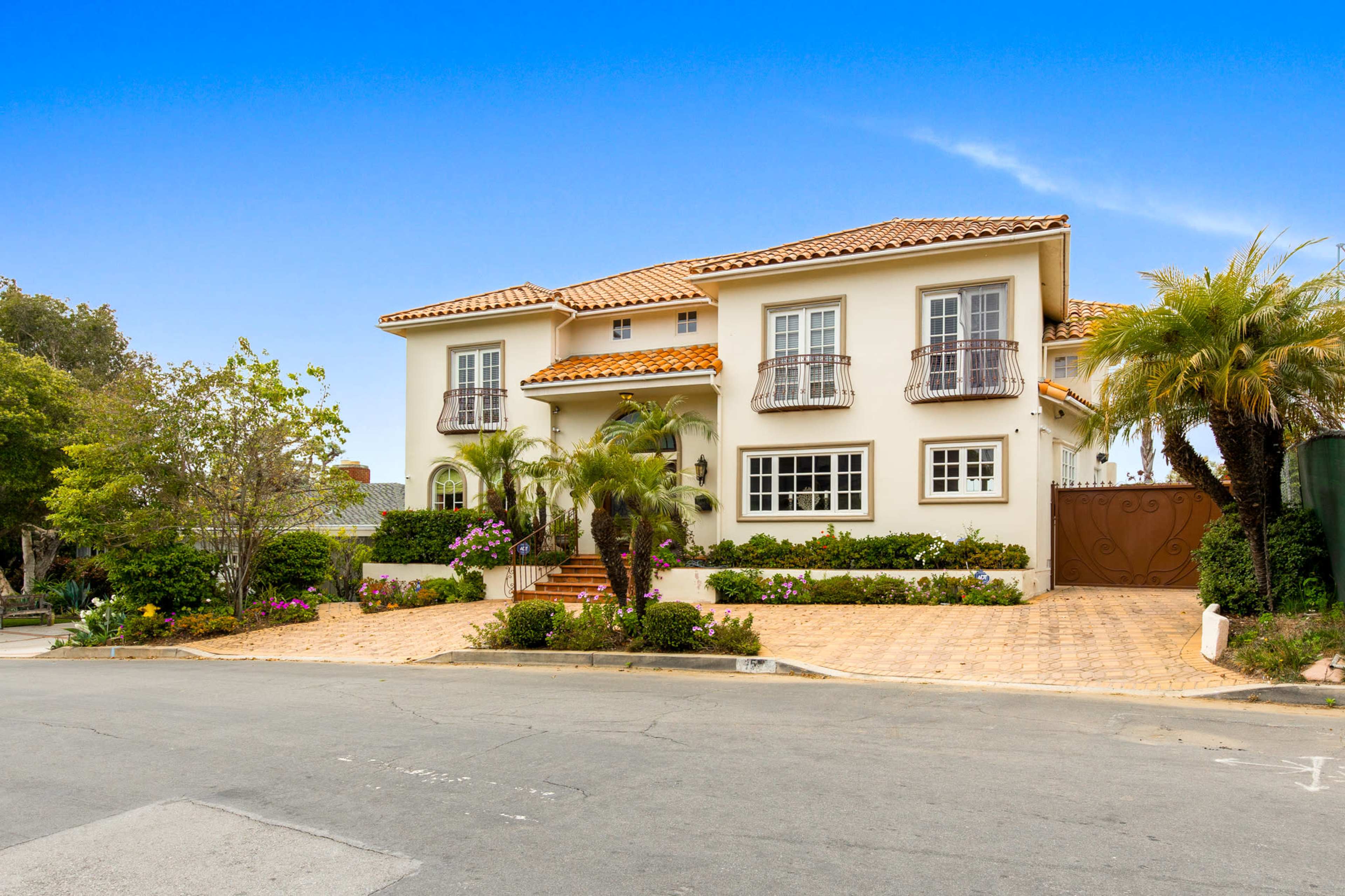 A two-story house with a tiled roof and balcony features is situated at the corner of a street, surrounded by palm trees and decorative landscaping.
