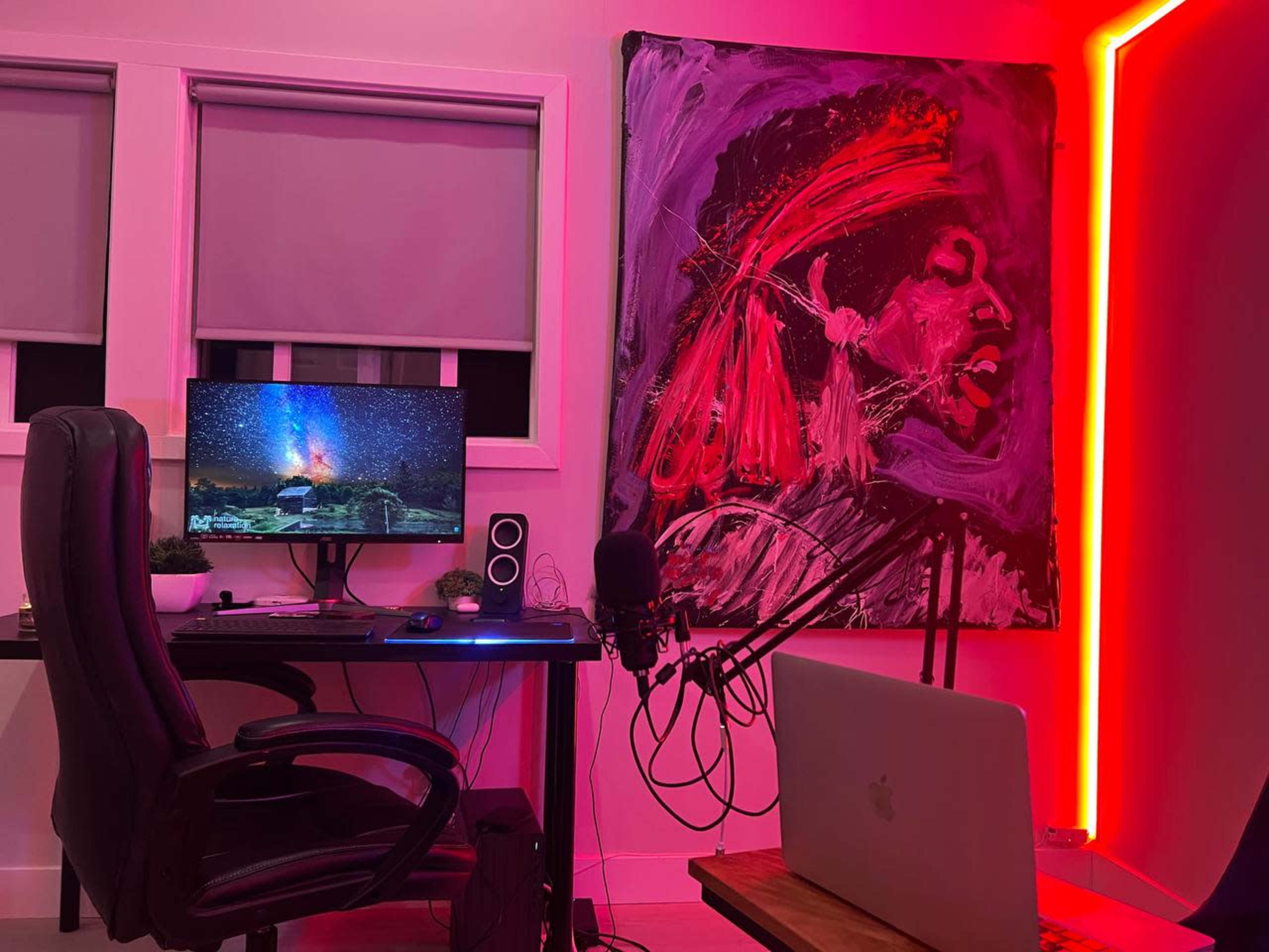 A modern home office setup featuring a desk with a computer monitor, a microphone, and a laptop, alongside a colorful painting and illuminated walls.