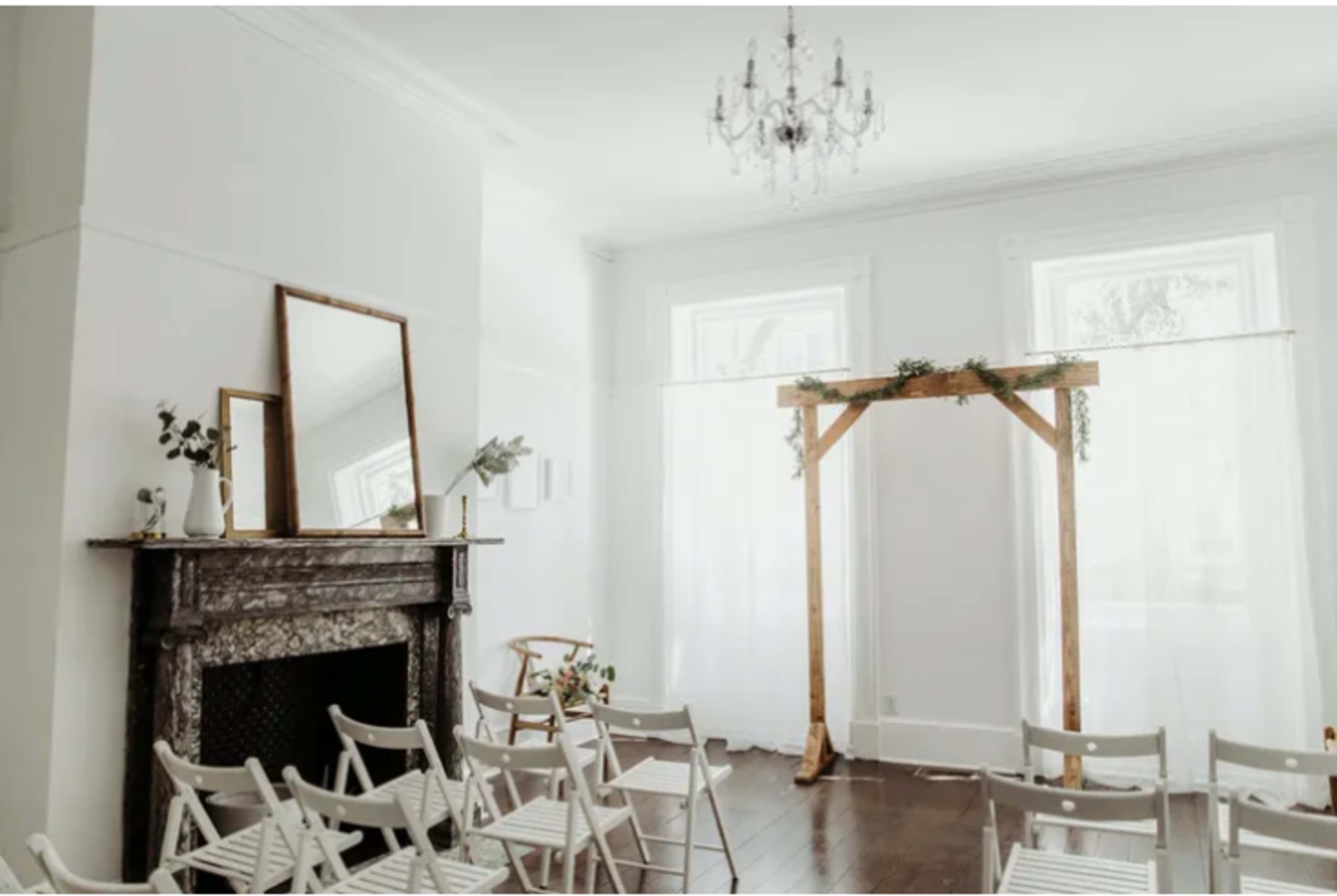 The room features a simple fireplace, a large mirror, and a wooden arch decorated with greenery, surrounded by white folding chairs.