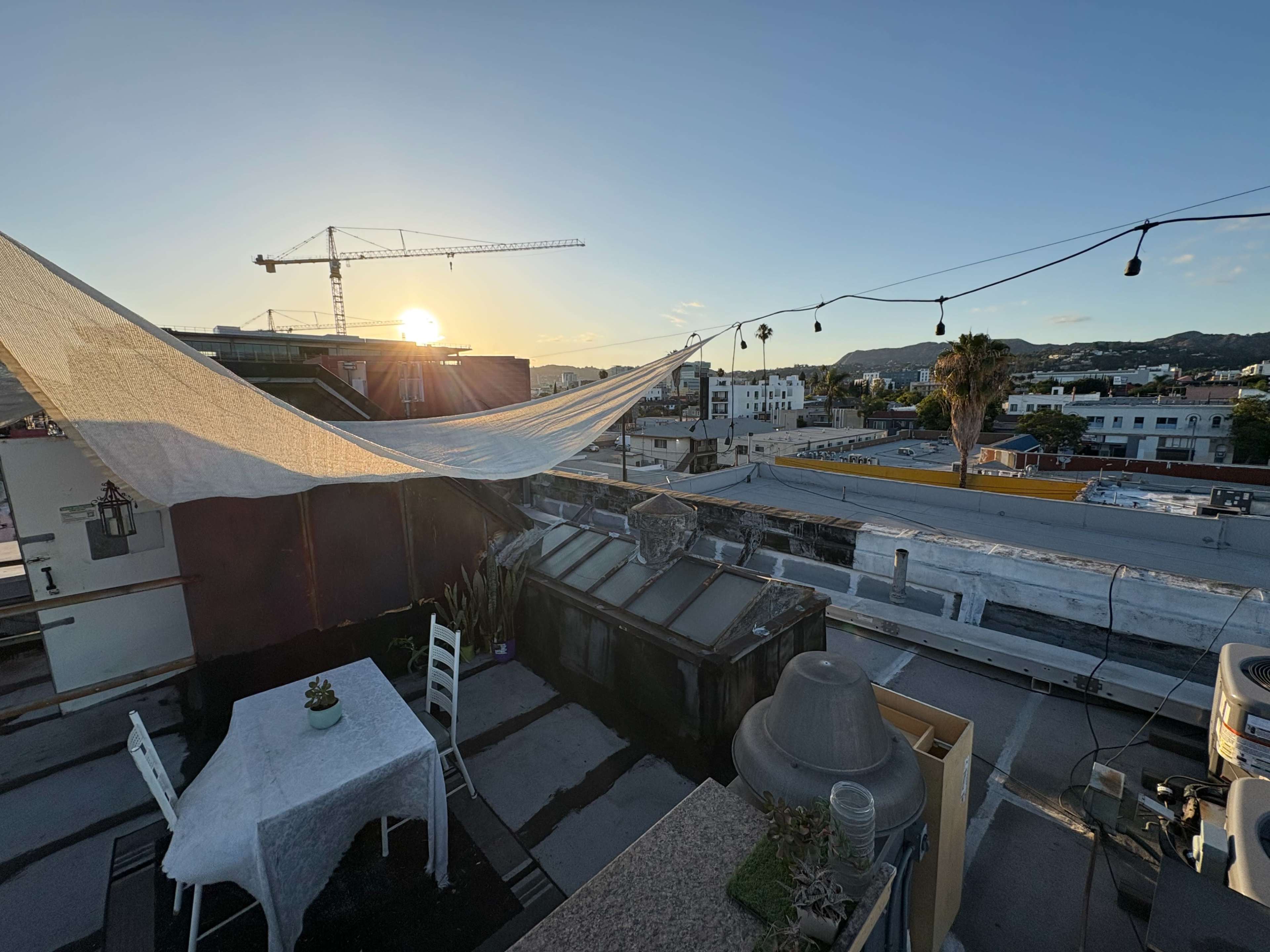 The image shows a rooftop terrace with a table, white tablecloth, and a large canopy, overlooking a city skyline at sunset.