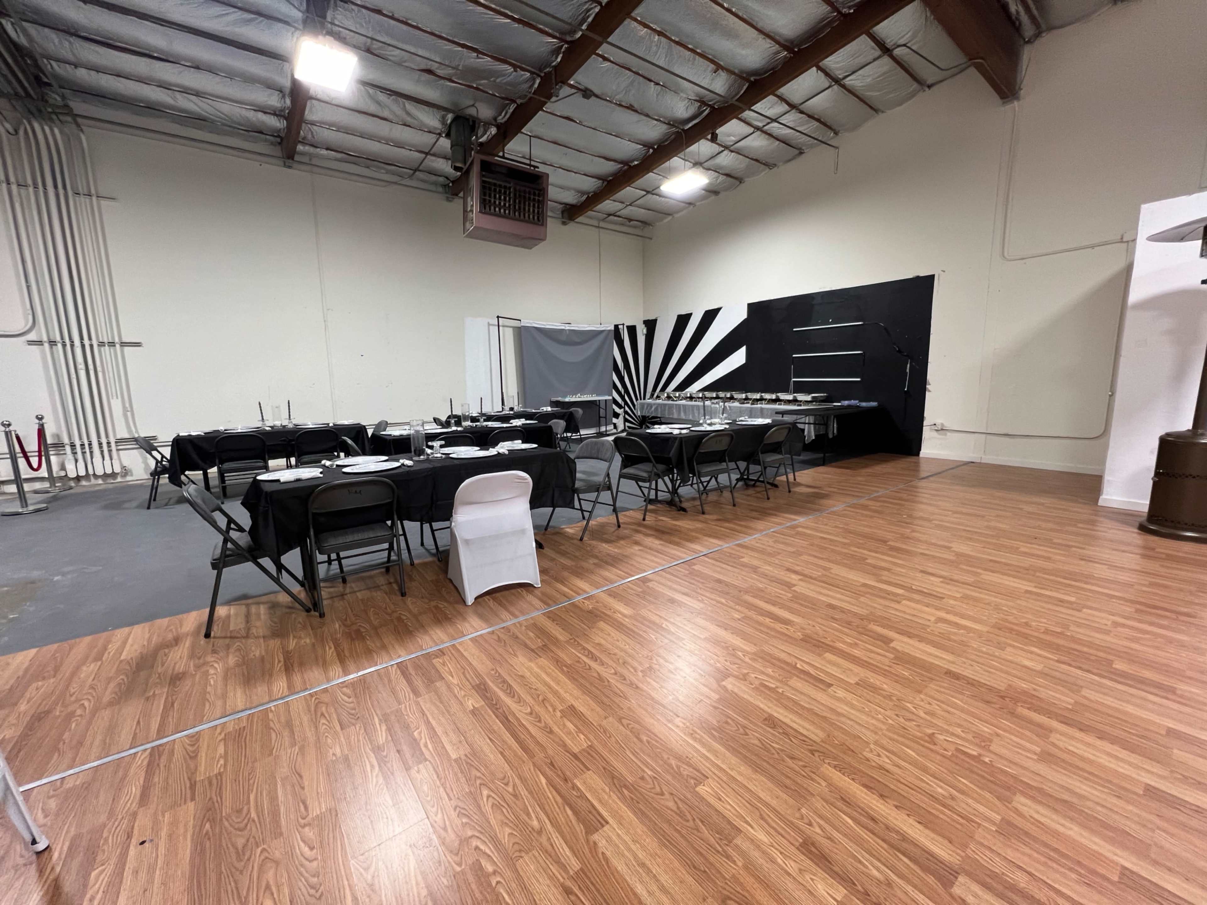 The image shows a spacious indoor area with a set of tables and chairs arranged for dining, featuring a black-and-white themed backdrop and polished wooden flooring.