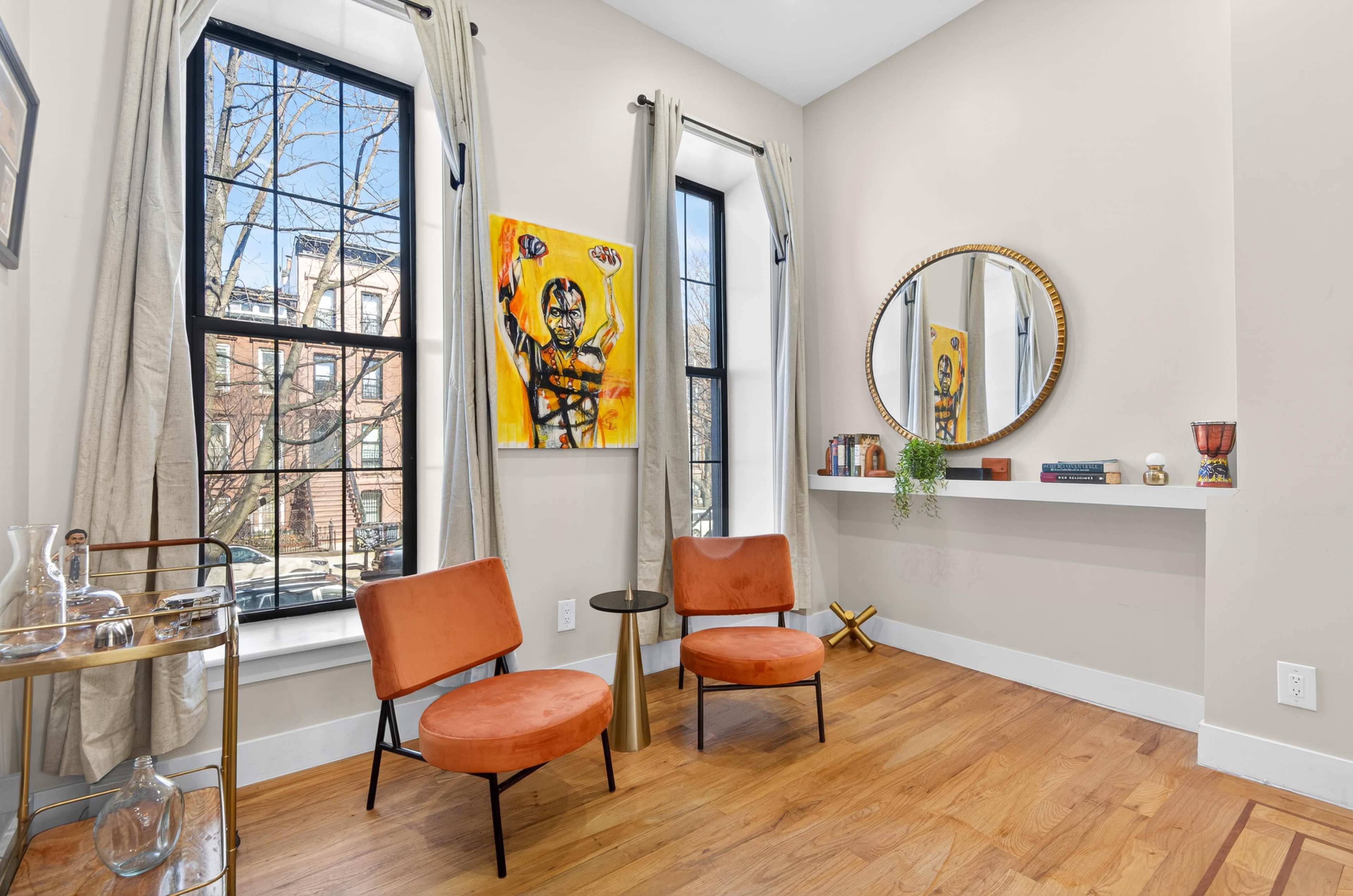 The image shows a small living area with two orange chairs, a round mirror, a wall-mounted shelf, and large windows letting in natural light.