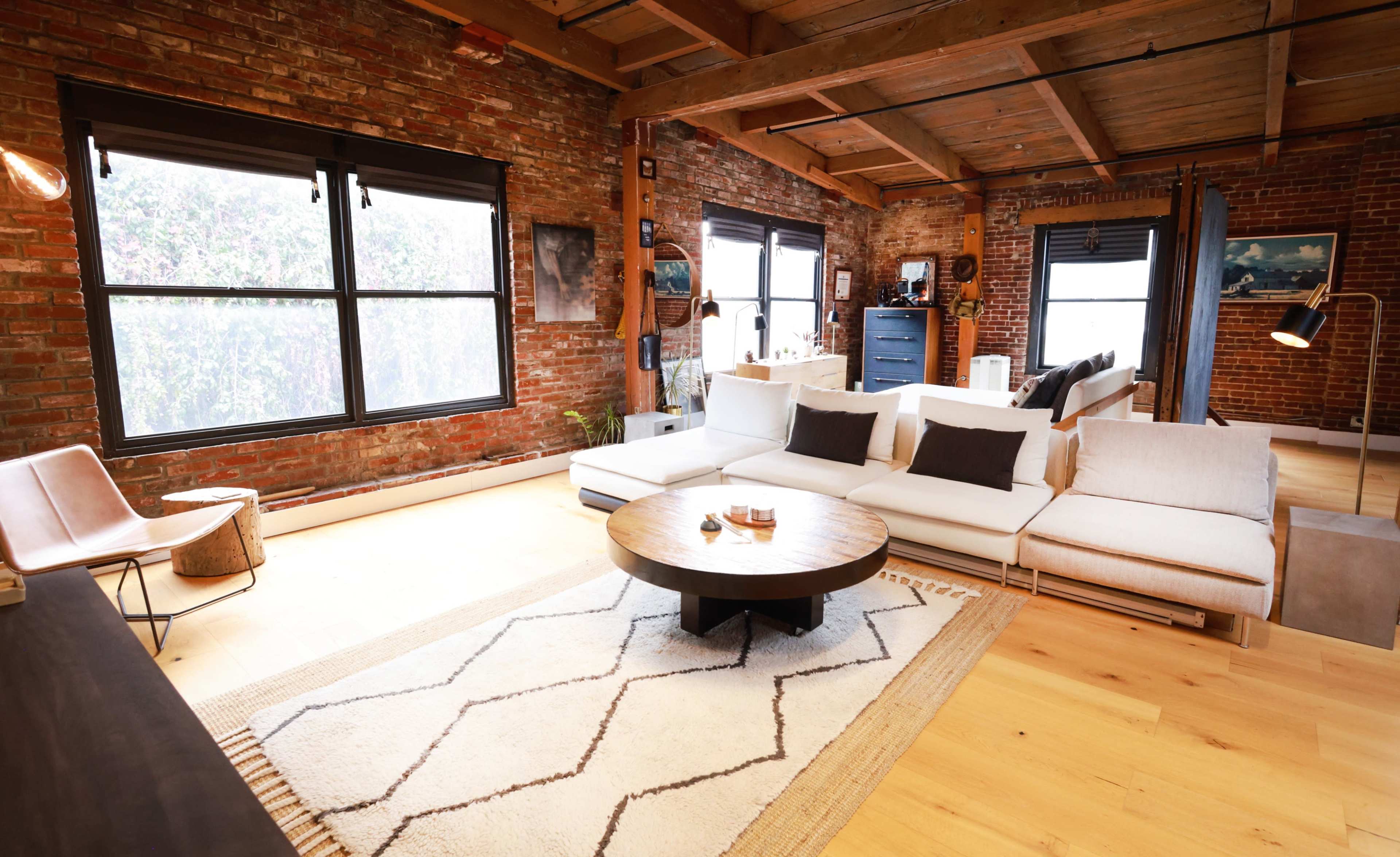 The image shows a modern living room with exposed brick walls, large windows, a sectional sofa, a round coffee table, and light wooden flooring.