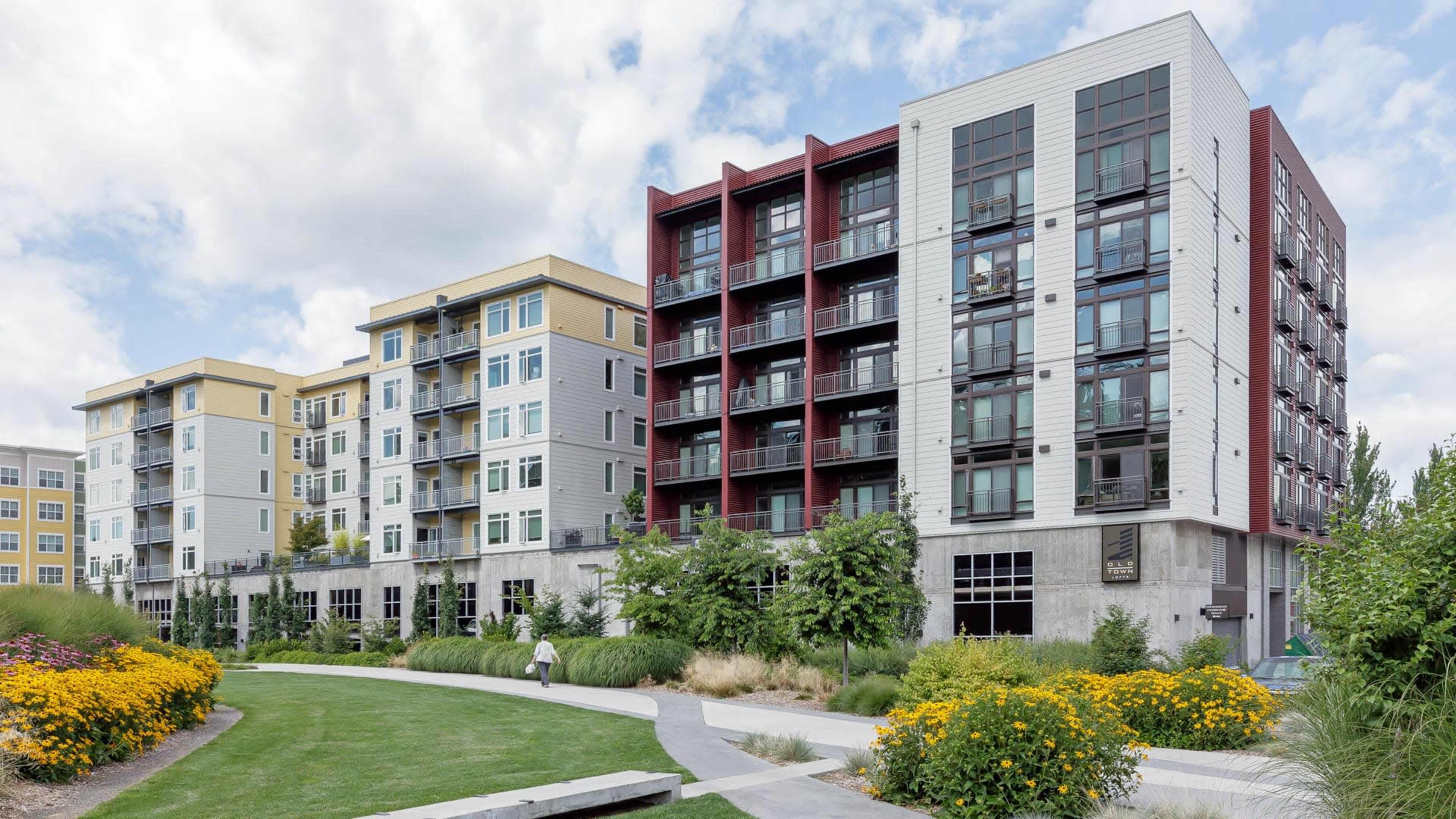 A row of modern apartment buildings with balconies and landscaped gardens are set alongside a paved walking path.