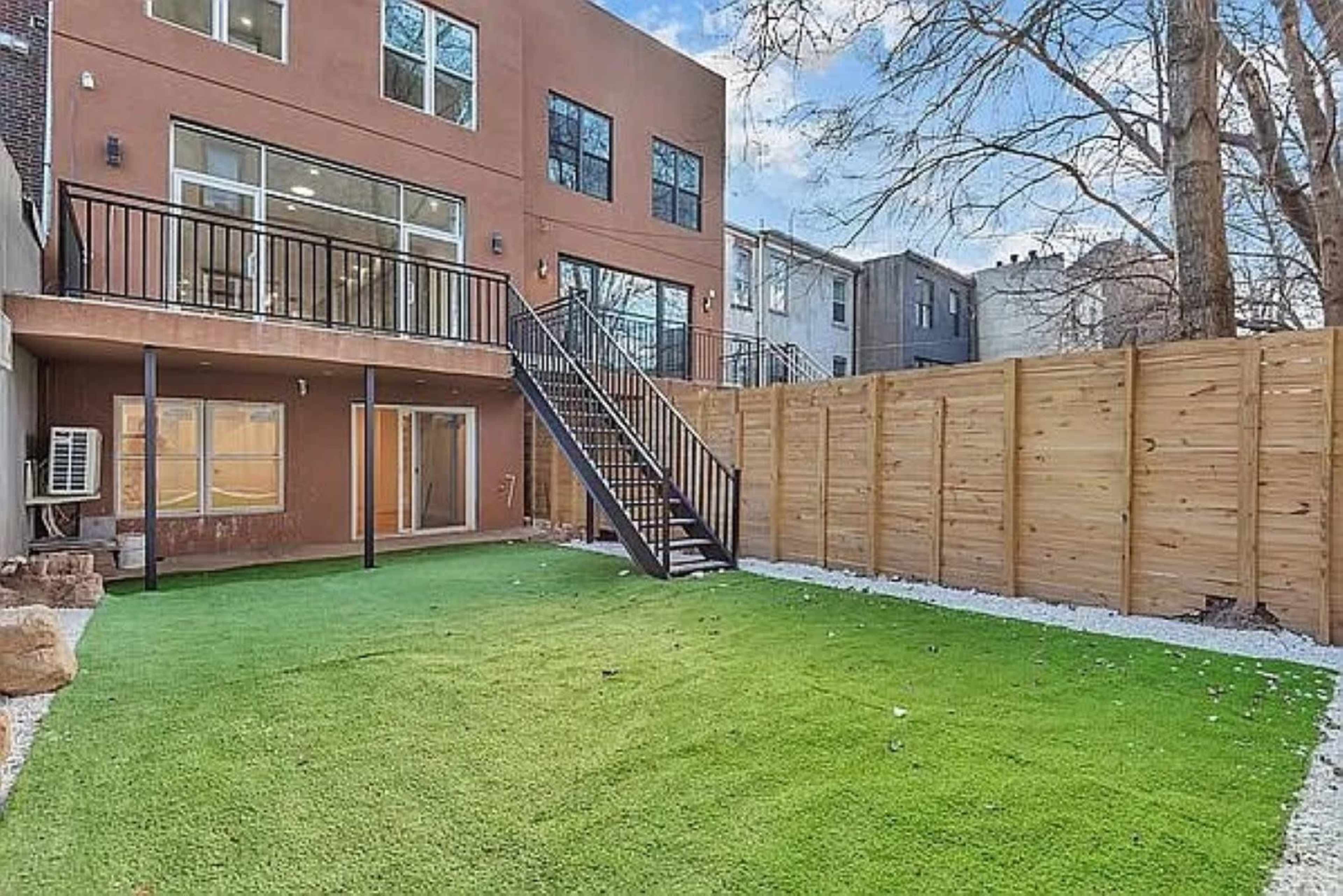 The image shows a backyard with artificial grass, wooden fencing, and a staircase leading to an upper floor of a two-story building.