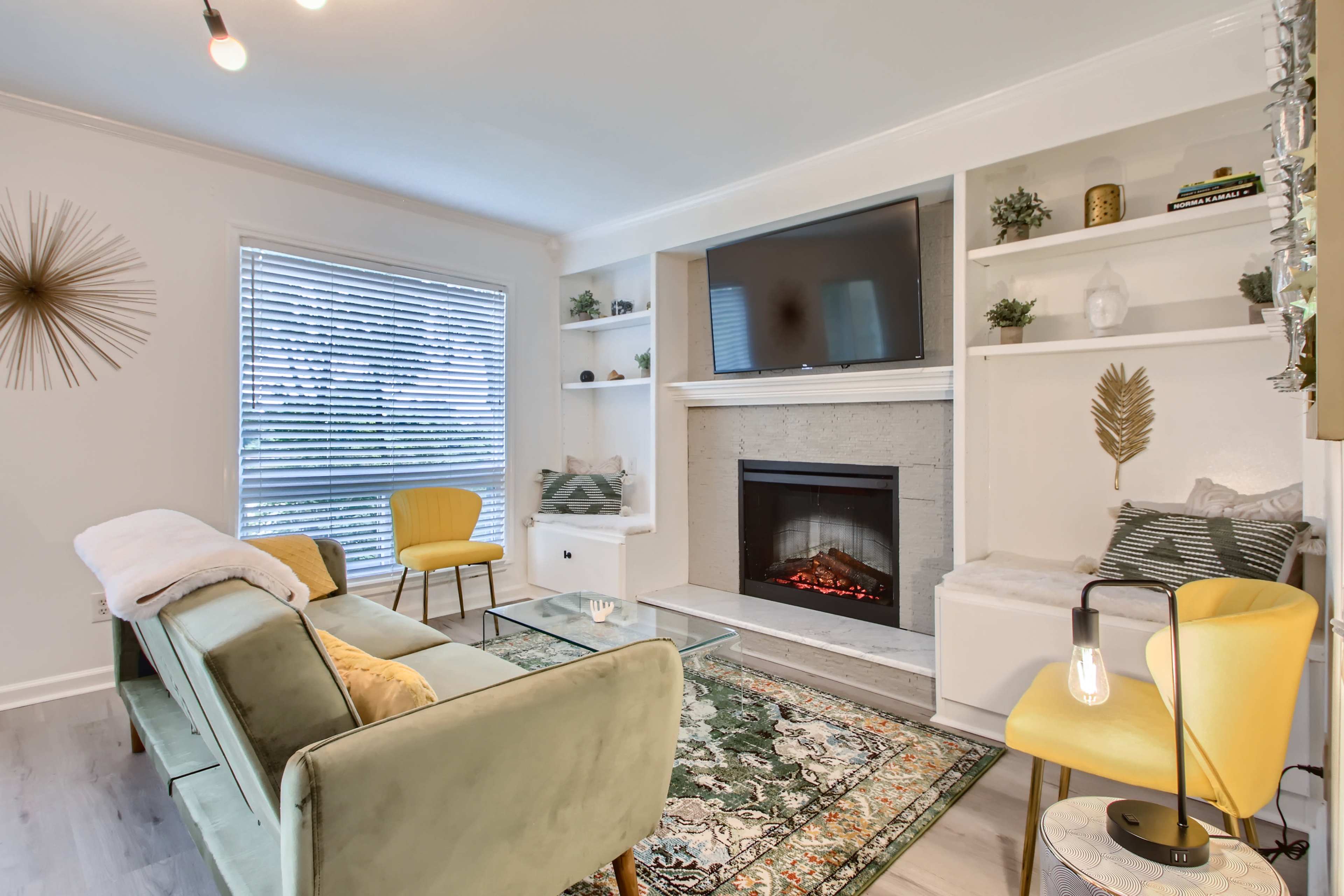 A modern living room featuring a cozy fireplace, a television above it, bookshelves on either side, and a pair of yellow chairs surrounded by a green sofa and a patterned area rug.