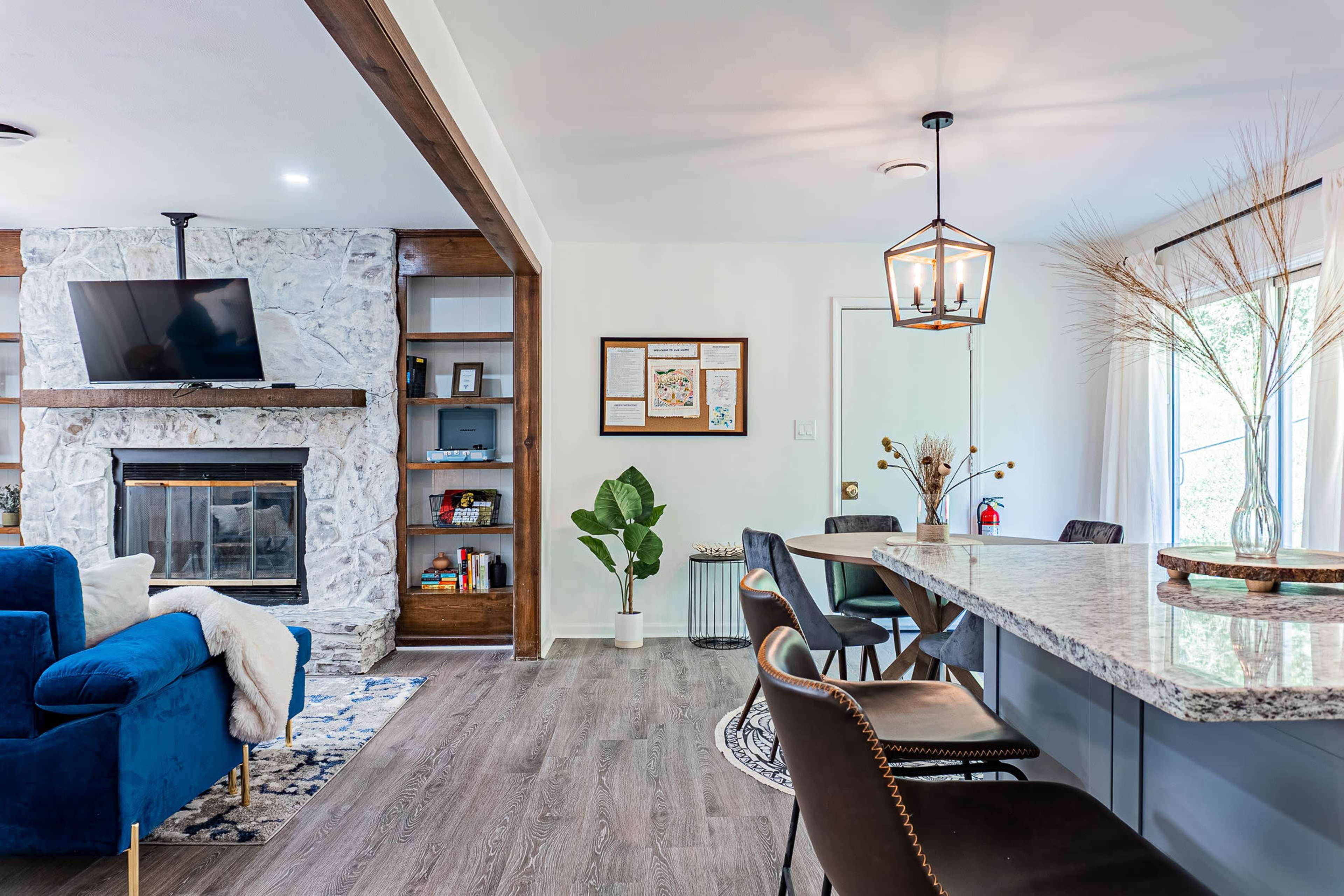 A modern living space featuring a blue sofa, a stone fireplace with a mounted TV, a dining area with a large granite table, and a potted plant.