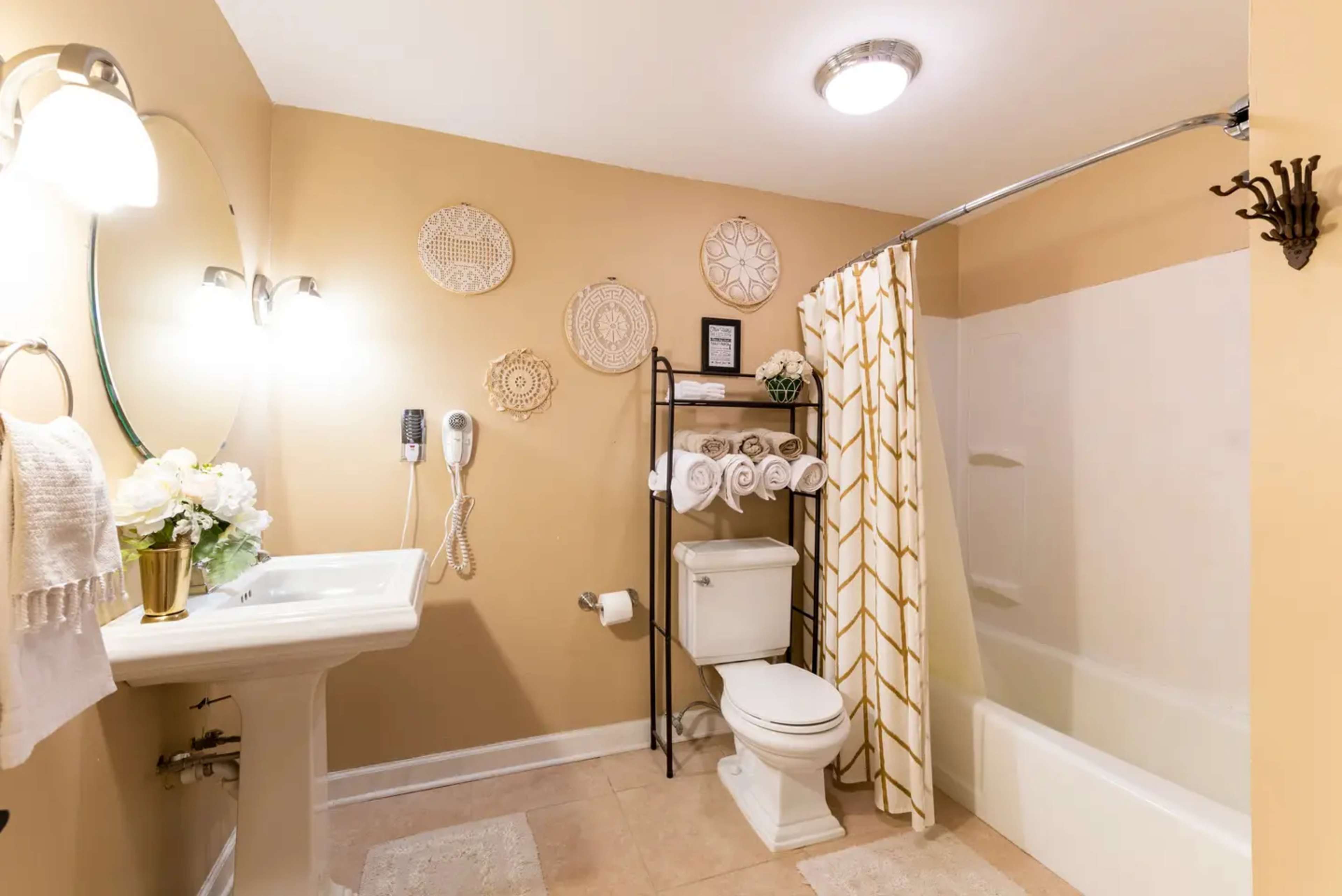 The bathroom features a sink with a mirror, a toilet, a bathtub with a shower curtain, and a shelving unit displaying neatly rolled towels.