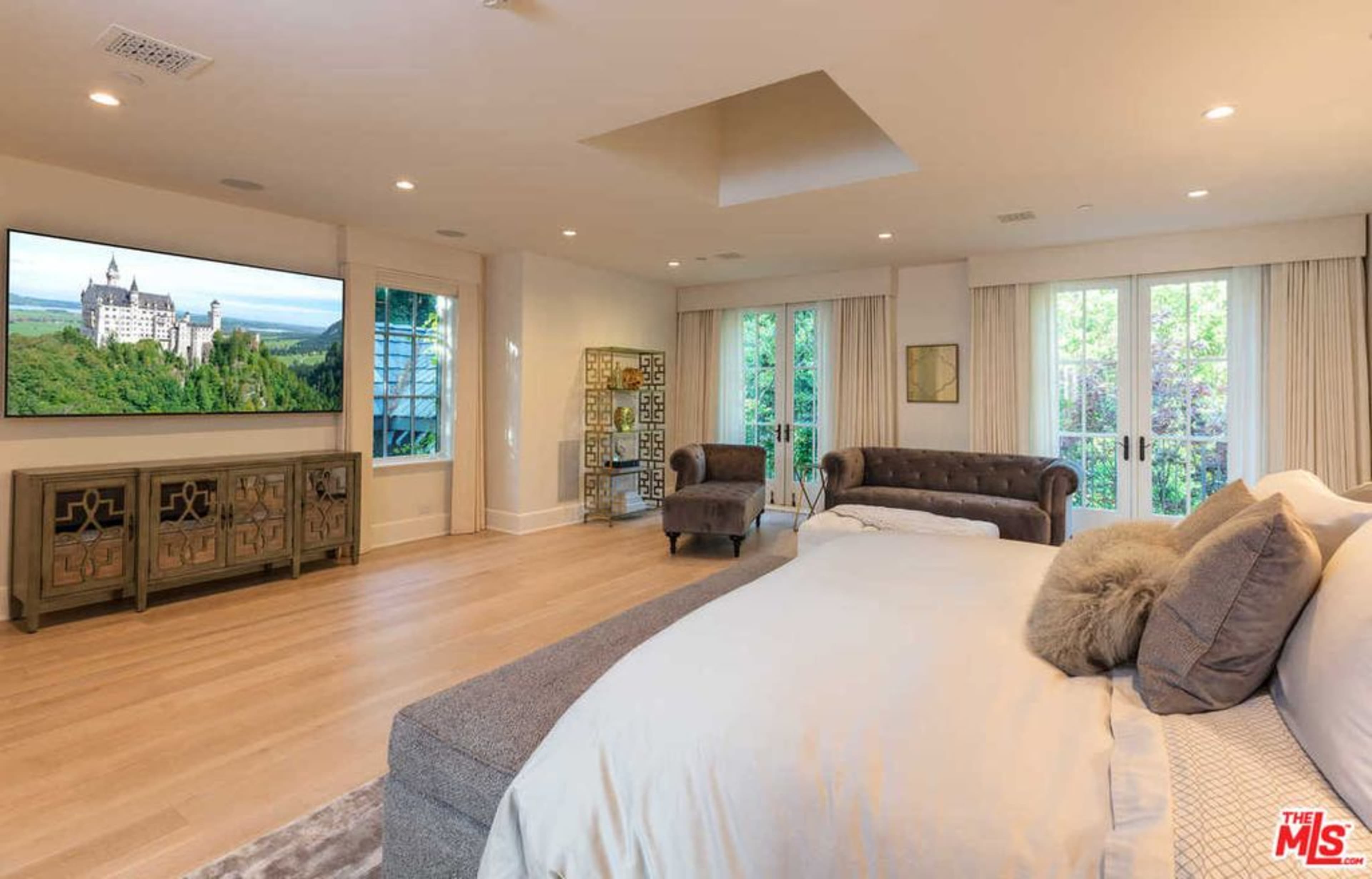 The image shows a spacious bedroom with a large bed, a tufted armchair, and a mounted flat-screen television displaying a castle landscape.
