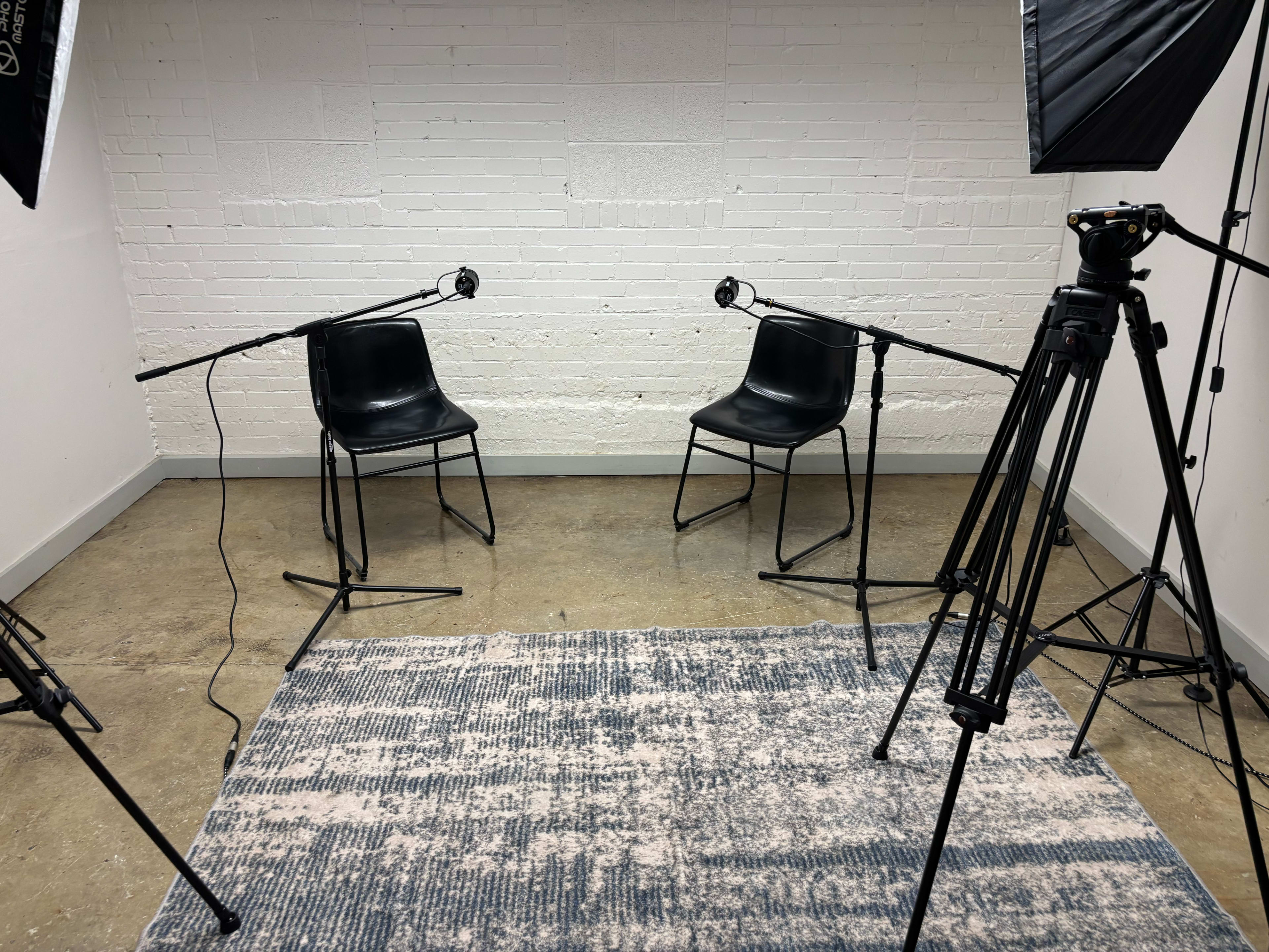 The image shows a room with two black chairs facing each other, each equipped with a microphone, and surrounded by lighting equipment on tripods.