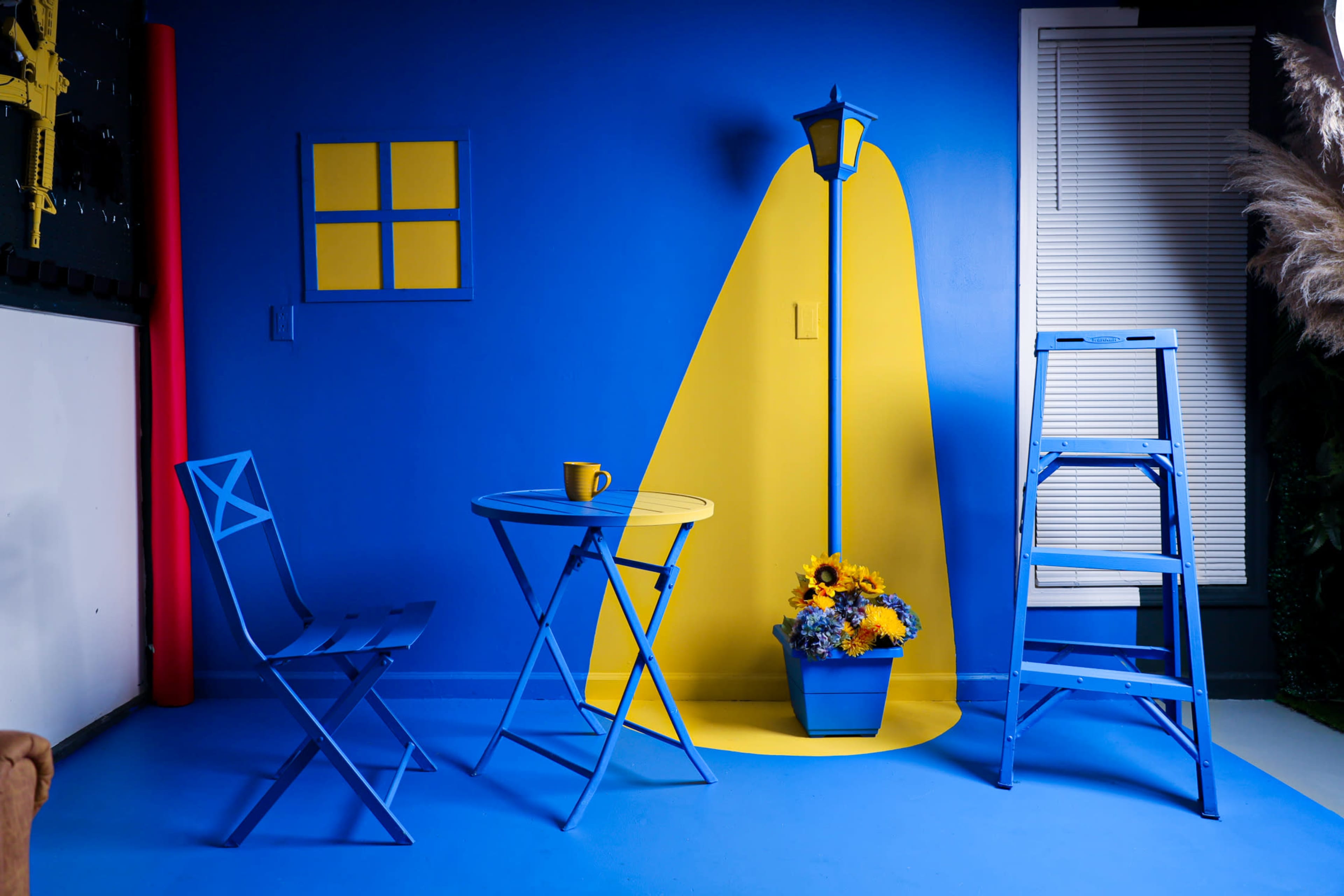 A blue room with a yellow accent wall, a small round table and chair set, a yellow lamp post with a flower pot, and a step ladder, all arranged against a vibrant blue backdrop.