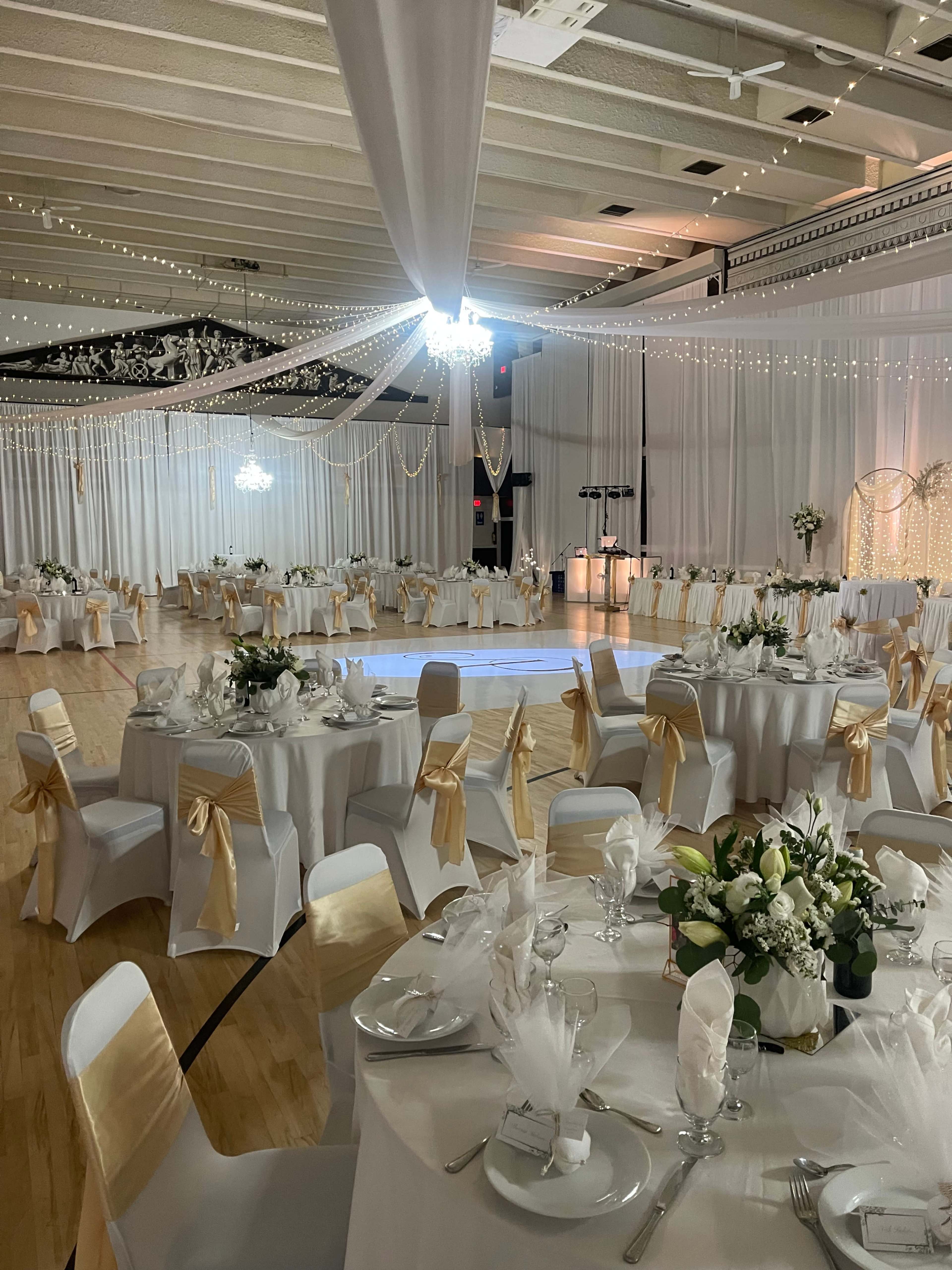 The image shows a decorated indoor venue with tables set for a formal event, featuring white tablecloths and gold accents, illuminated by soft lighting and draped fabric.