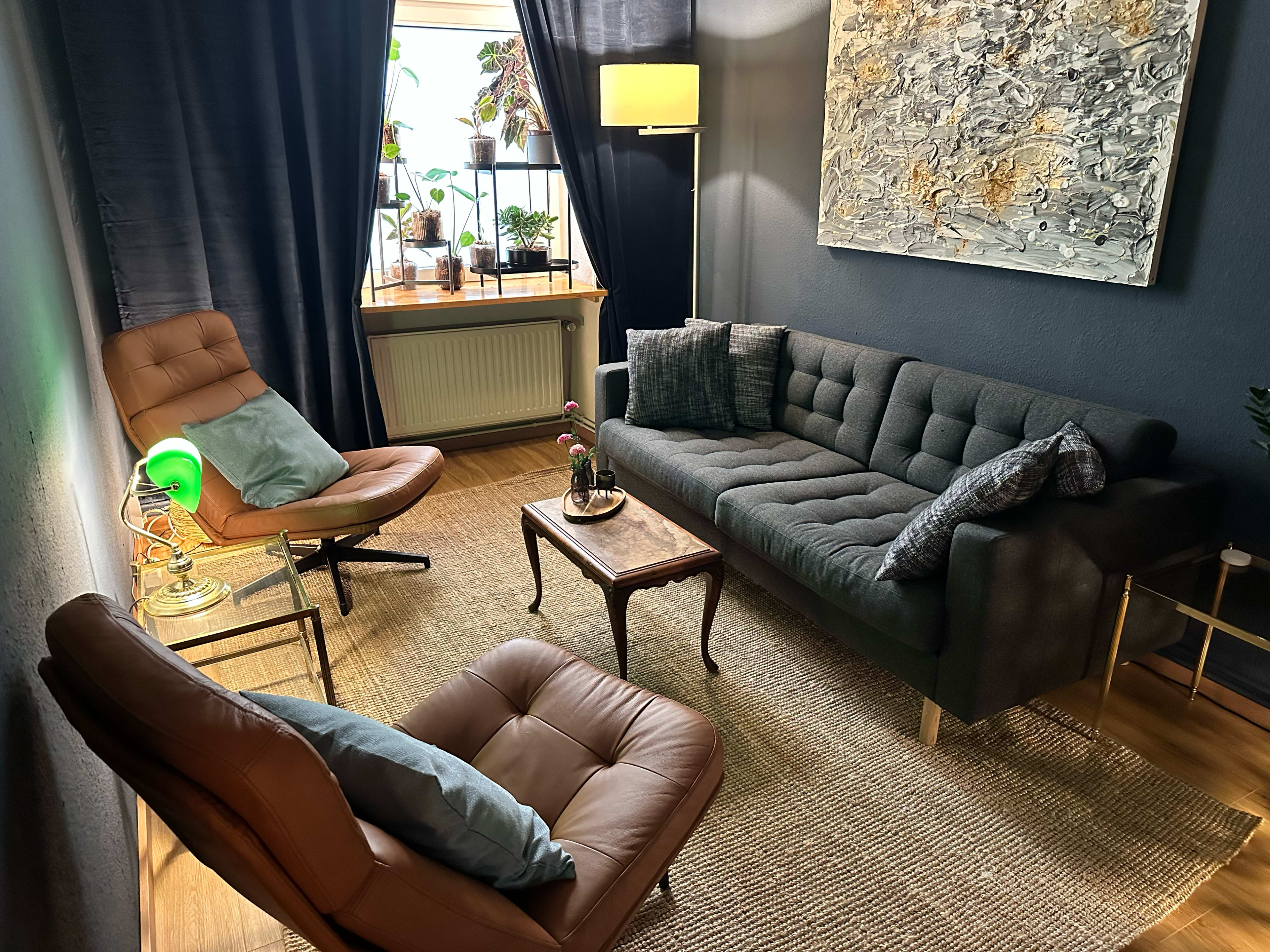 The image shows a cozy living room with a dark color scheme, featuring a modern sofa, a pair of accent chairs, and a decorative coffee table, all illuminated by natural light from a window surrounded by plants.