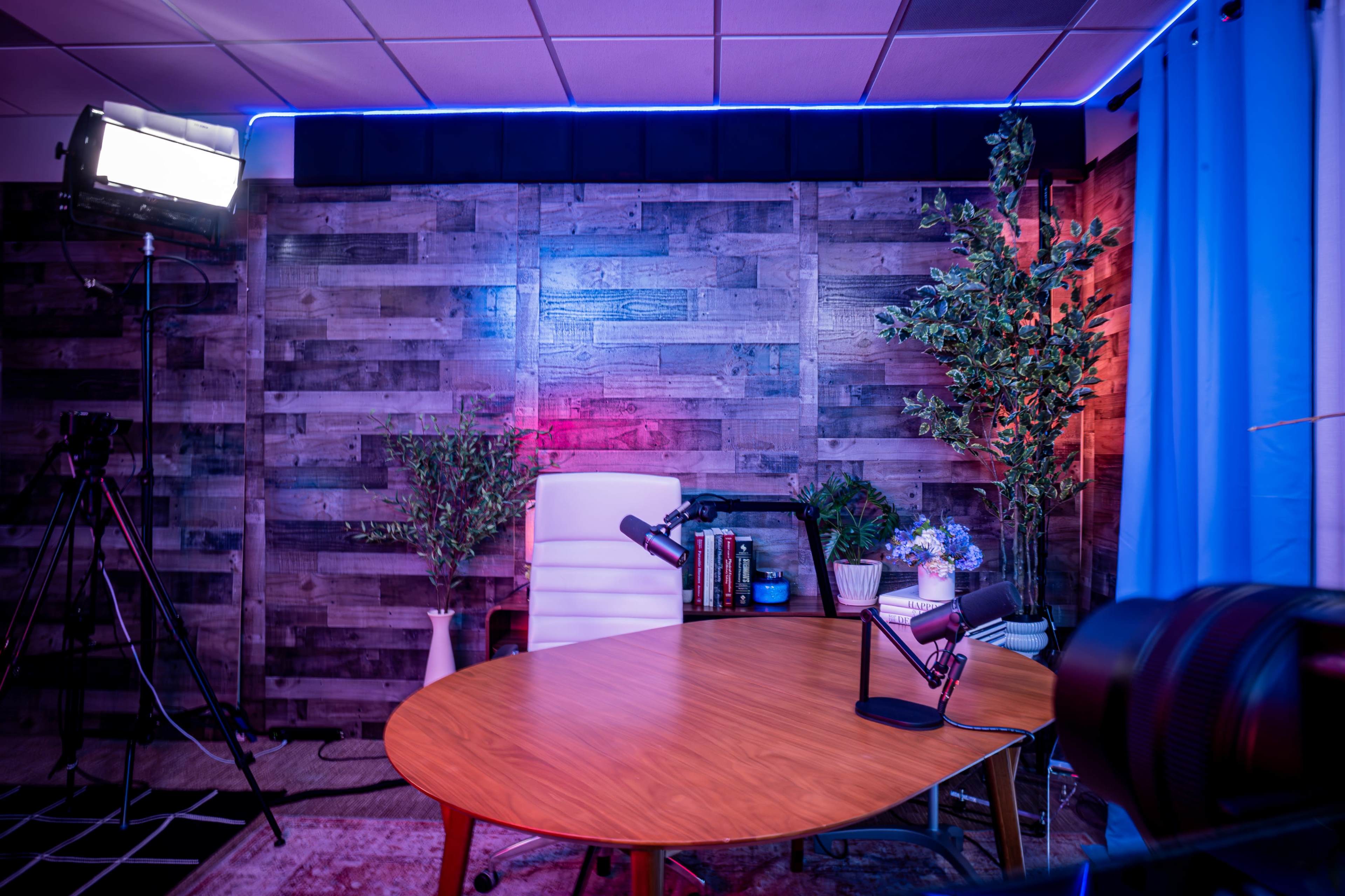 The image shows a modern podcast studio featuring a wooden table, a white chair, plants, and professional lighting against a textured wall.