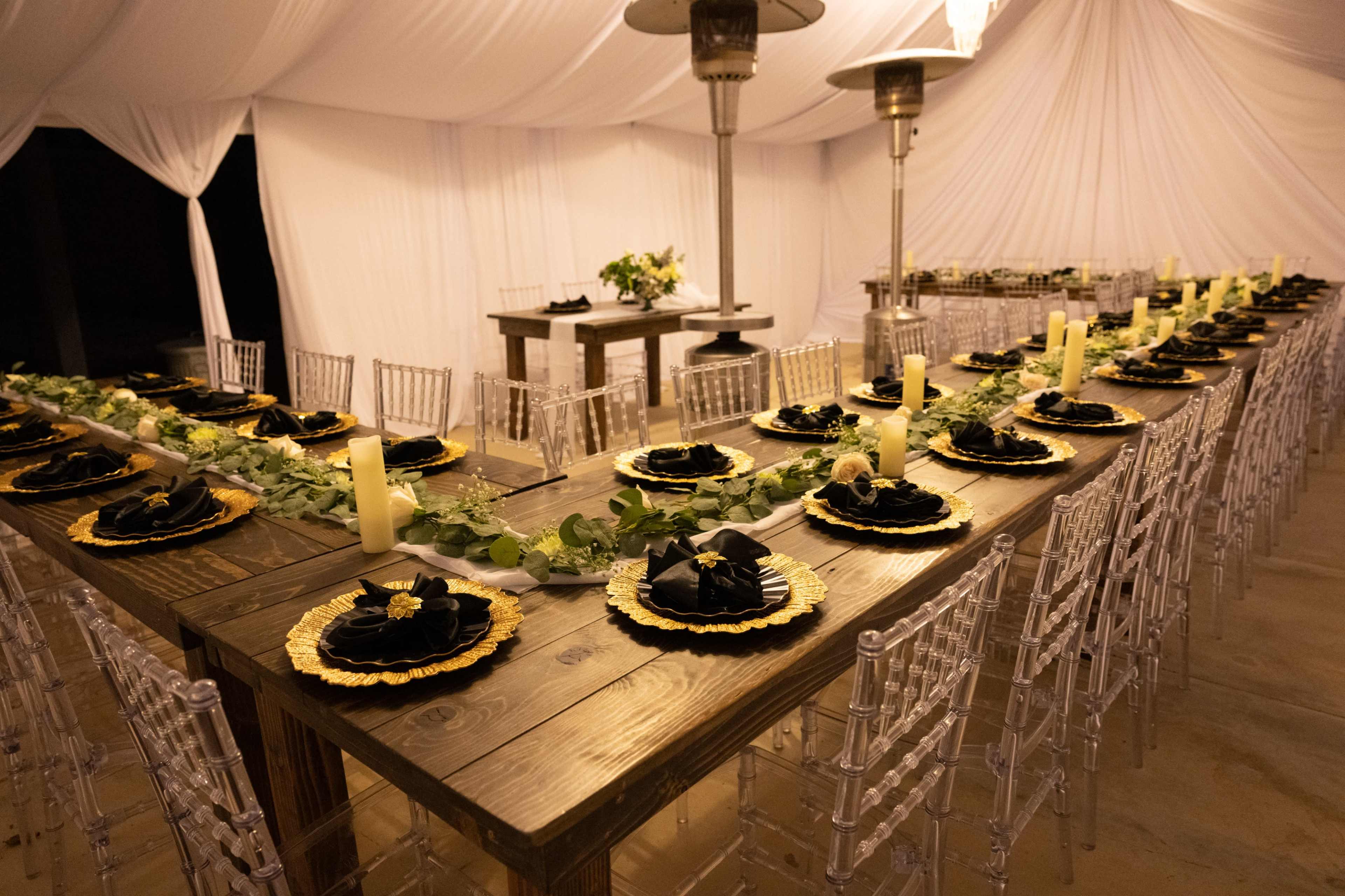 A long wooden table is elegantly set with black plates and gold accents, surrounded by clear plastic chairs and decorated with greenery, in a tented space with white drapery.