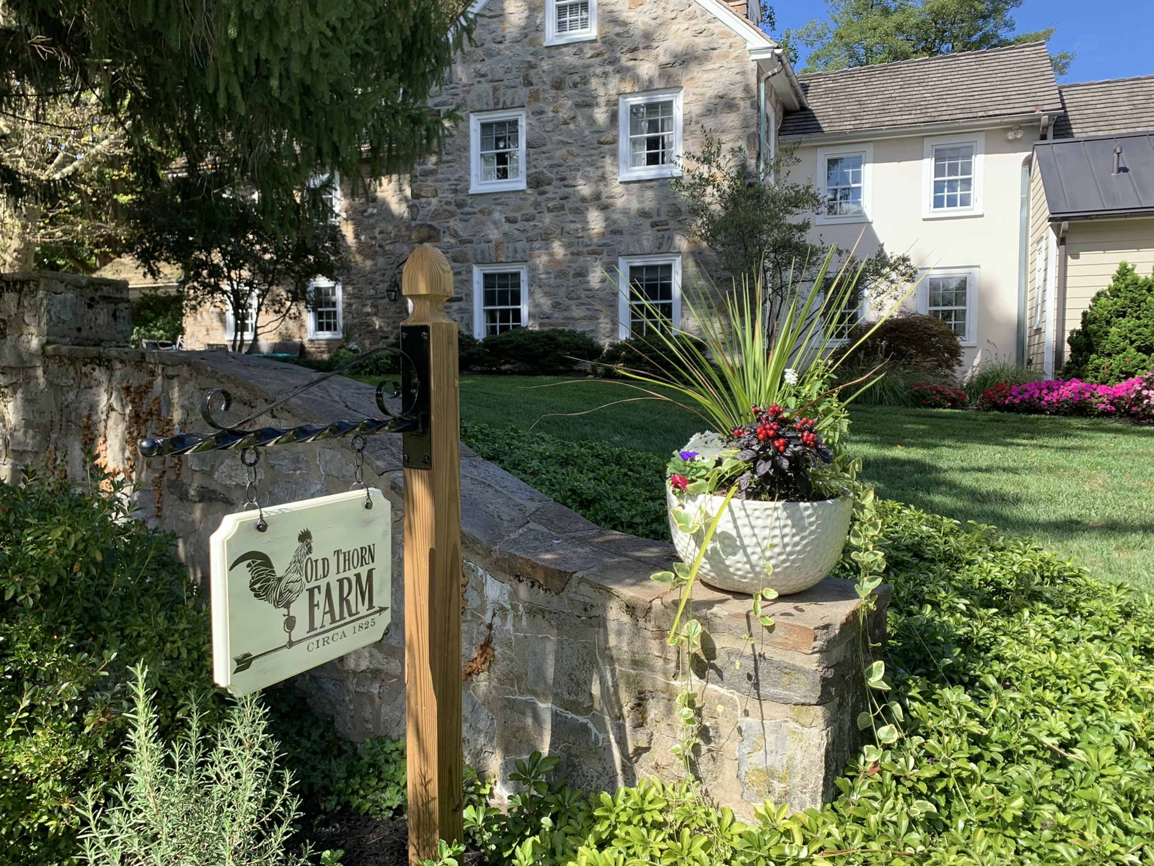 A stone wall and sign for "Old Thorn Farm," with a well-kept house and colorful flower beds in the background.