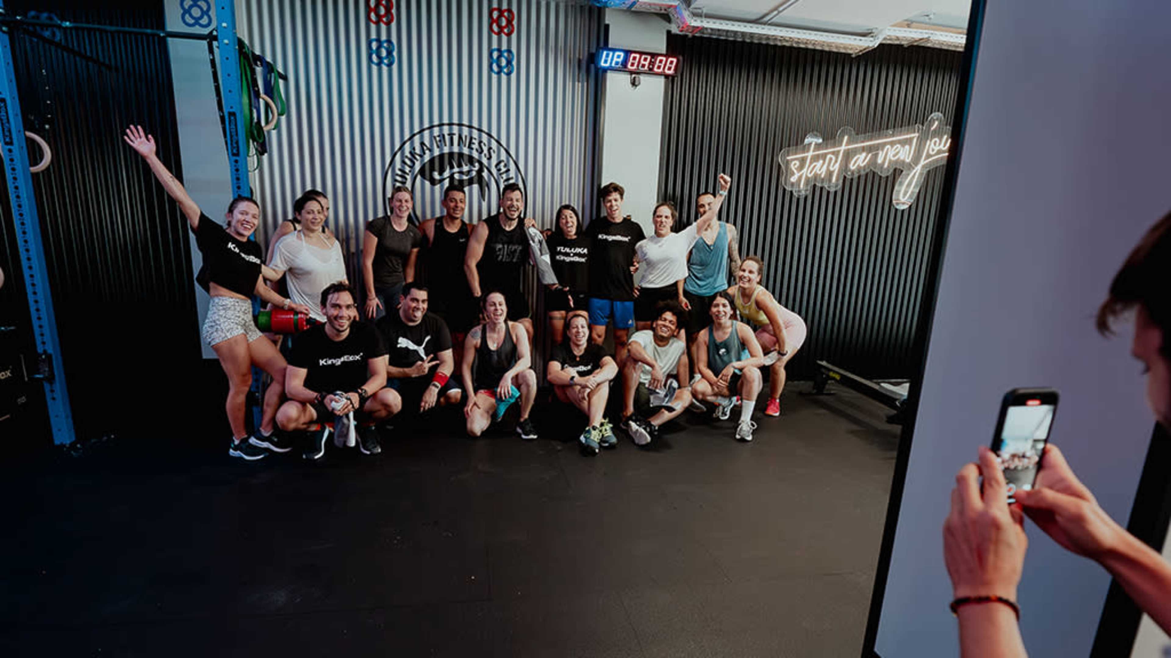 A group of people poses together in a gym after a fitness class, with some smiling and others holding equipment.