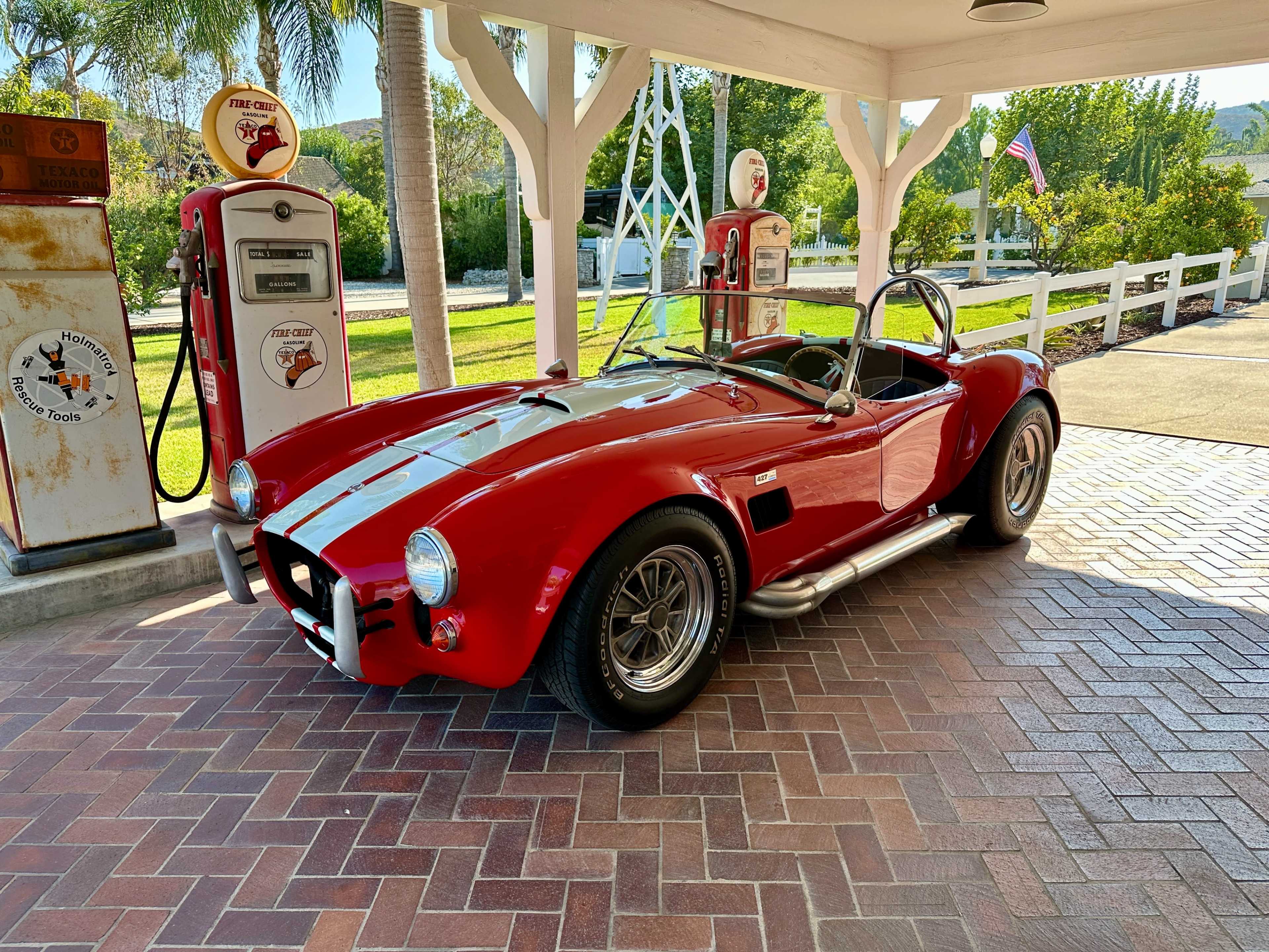 A red sports car with a black stripe is parked under a canopy beside vintage gas pumps.