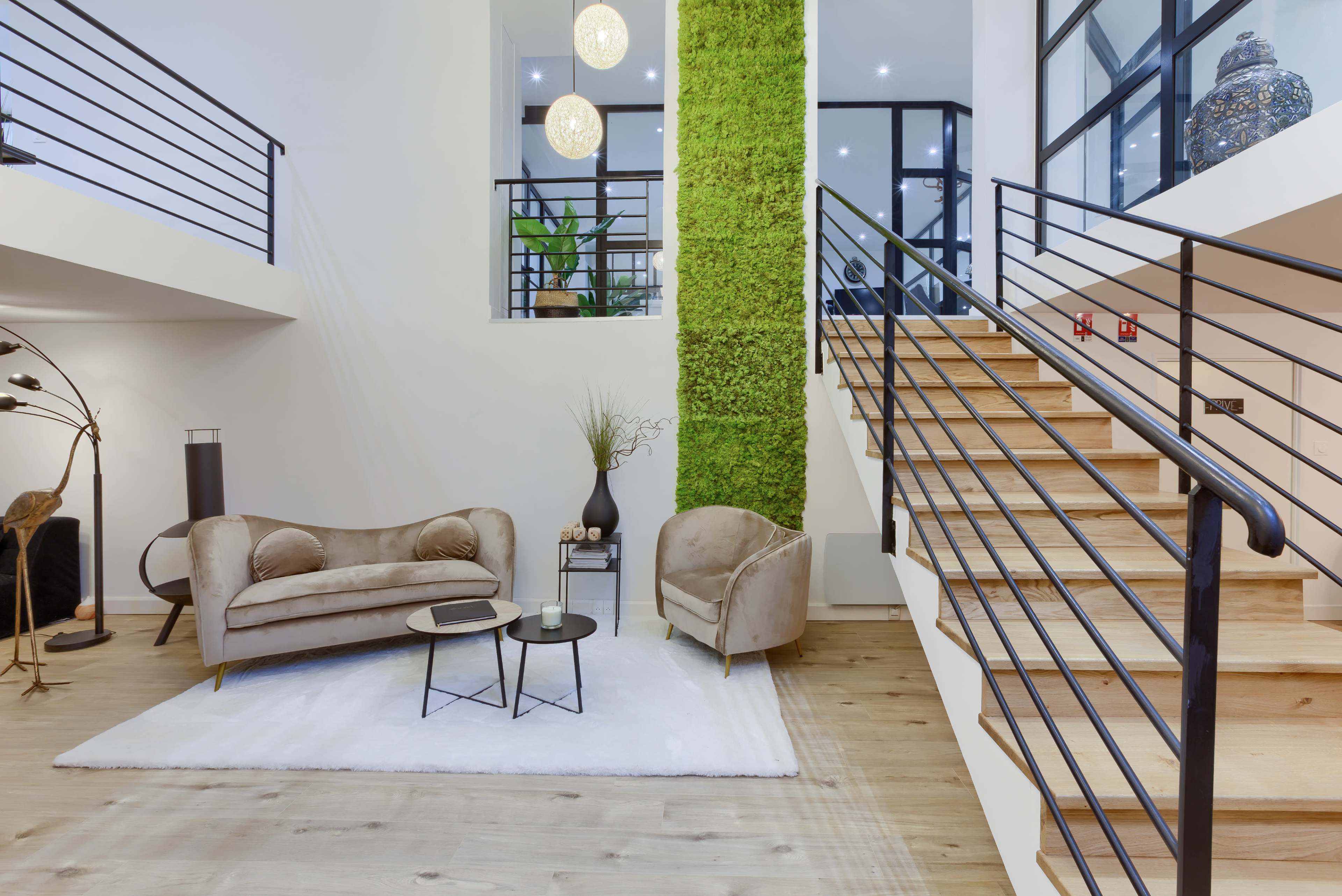 A modern living space with a large staircase, a green moss wall, a cozy seating area, and stylish lighting fixtures.