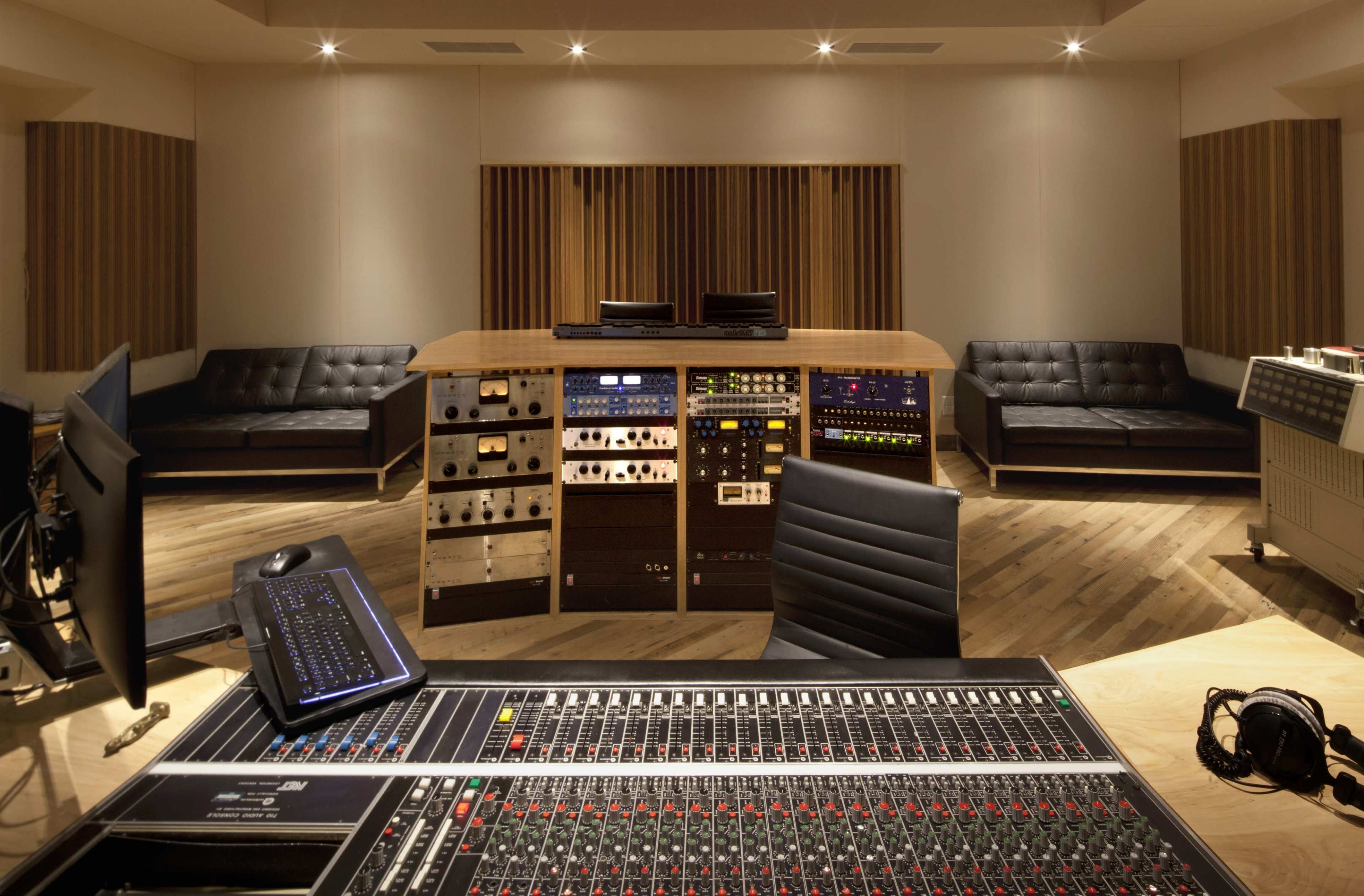 The image shows a modern recording studio with a central mixing console, sound equipment, and seating areas.