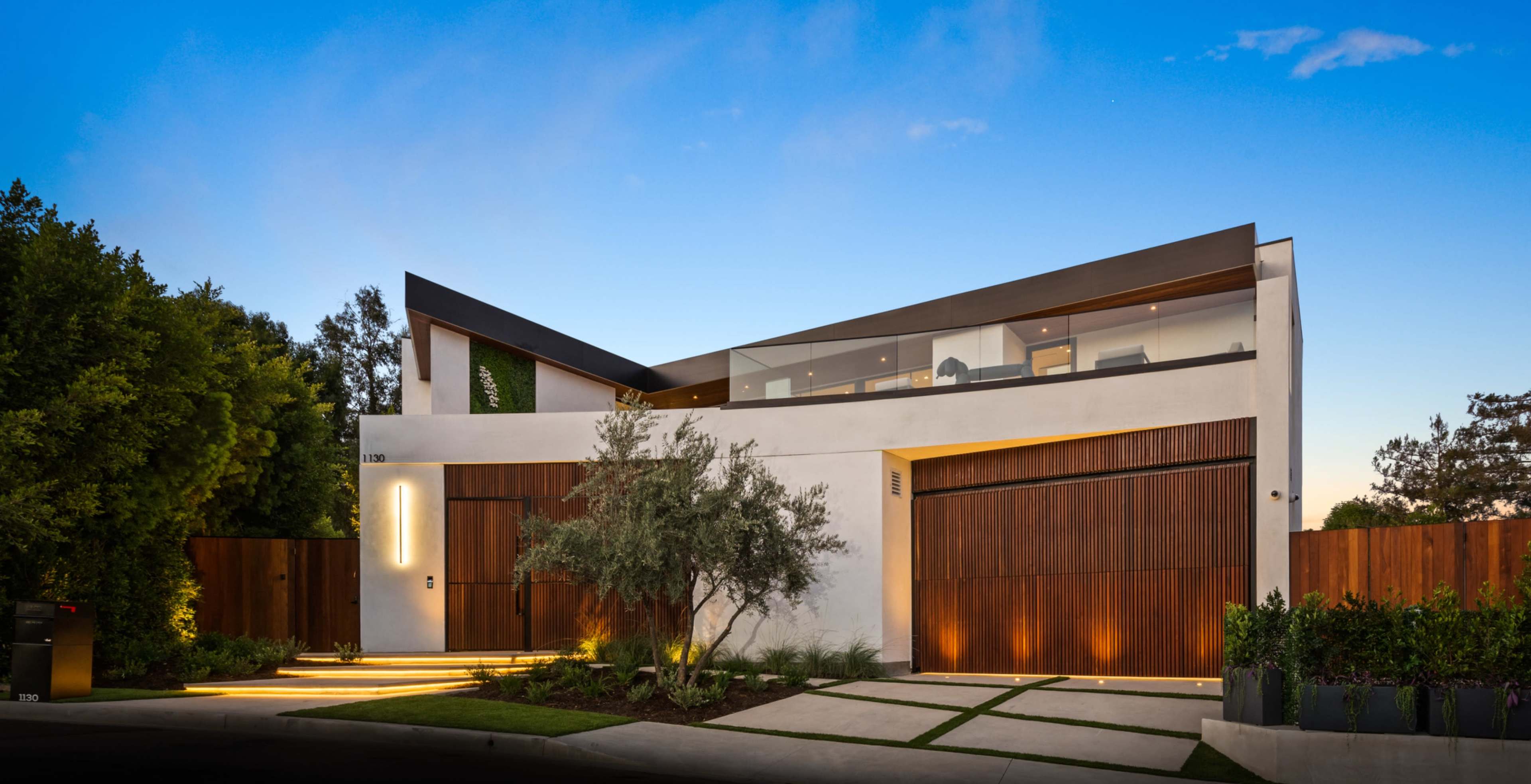 The image shows a modern two-story house with a flat roof, large windows, and wooden accents, surrounded by landscaped greenery.