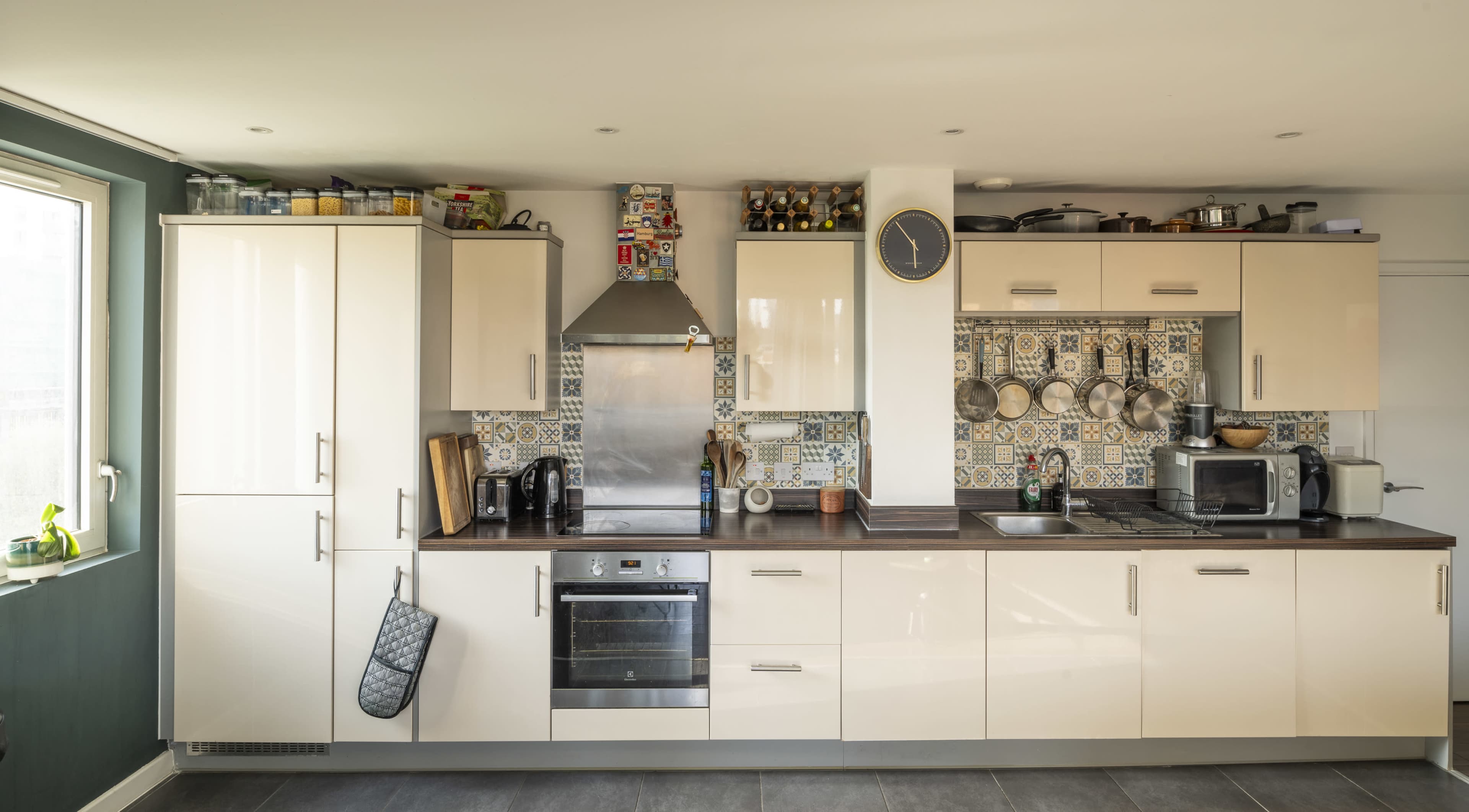 The image shows a modern kitchen with cream-colored cabinets, a stainless steel oven, and patterned tiles as a backsplash.